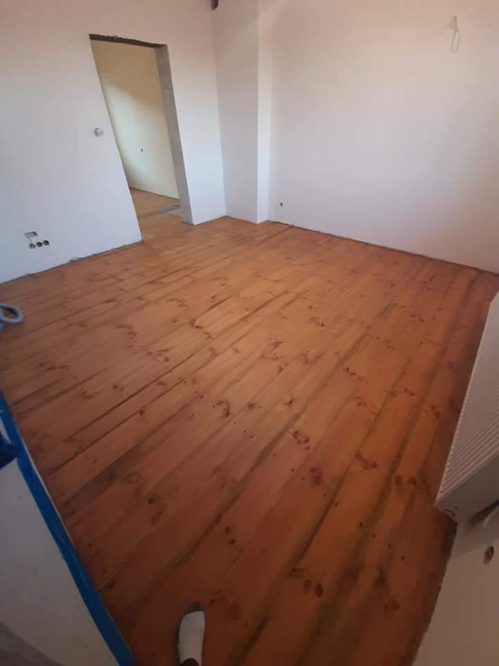 Wycyklinowana, drewniana podłoga w pokoju z surowymi ścianami, widoczne ślady po cyklinowaniu i fragment stopy w skarpetce.