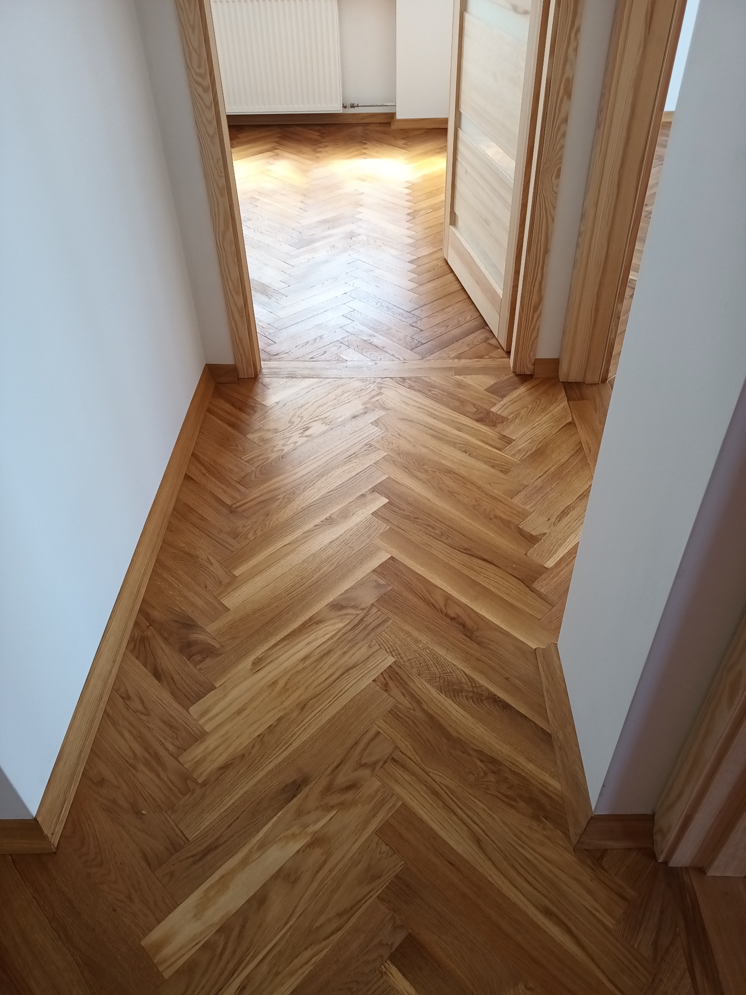 Ułożony parkiet w jodełkę w korytarzu, widoczne listwy przypodłogowe i ościeżnice z jasnego drewna, białe ściany, otwarte drzwi na końcu korytarza.