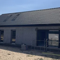 Niedokończony parterowy budynek z czarną dachówką, ocieplony szarą wełną mineralną, z wstawionymi oknami i drzwiami, na piaszczystym terenie.