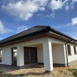 Parterowy dom modułowy z białymi ścianami i ciemną dachówką, drewniane okna, widok z zewnątrz w słoneczny dzień, teren jeszcze niezagospodarowany.