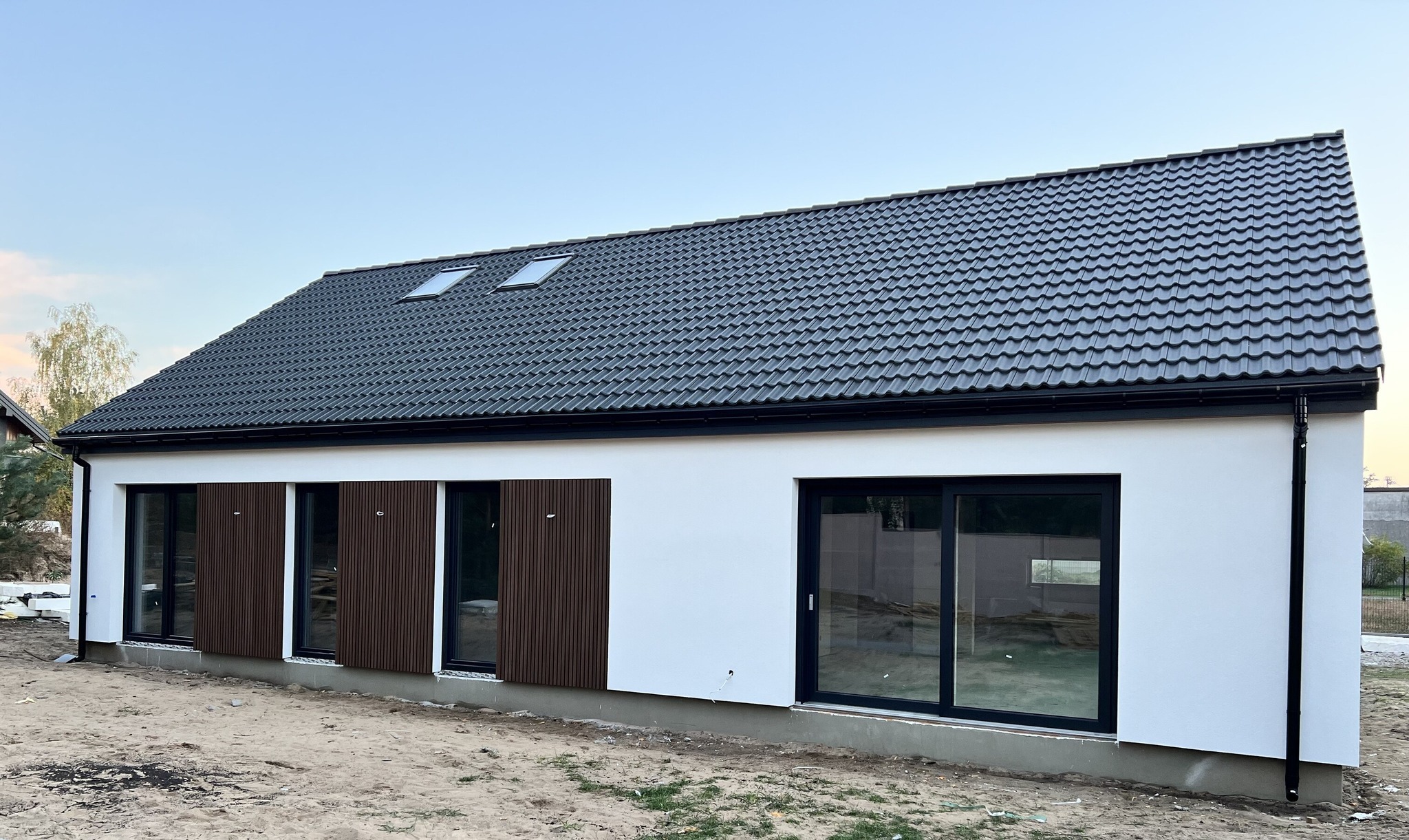Nowoczesny parterowy dom z białymi ścianami, grafitowym dachem z dachówki i brązowymi panelami dekoracyjnymi na elewacji, widoczne okna dachowe i przesuwne drzwi tarasowe.