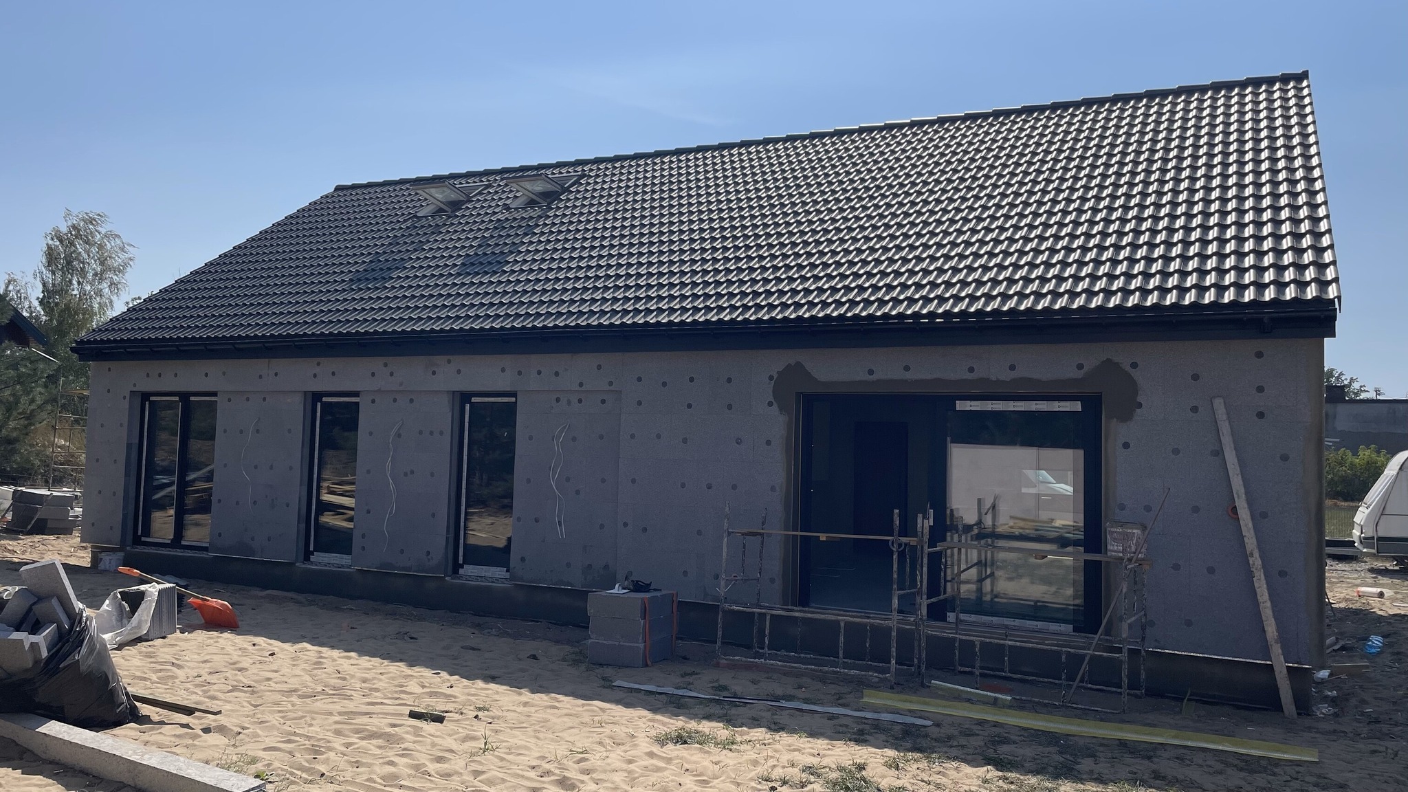 Niedokończony parterowy budynek z czarną dachówką, ocieplony szarą wełną mineralną, z wstawionymi oknami i drzwiami, na piaszczystym terenie.
