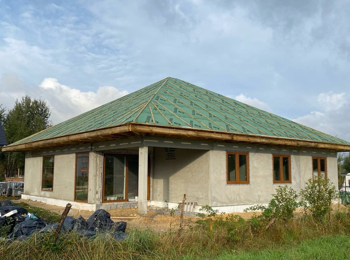 Surowy stan domu jednorodzinnego z keramzytu z widoczną konstrukcją dachu pokrytą zieloną membraną, brązowe okna, szare ściany zewnętrzne i sterta czarnych worków ze śmieciami na pierwszym planie.