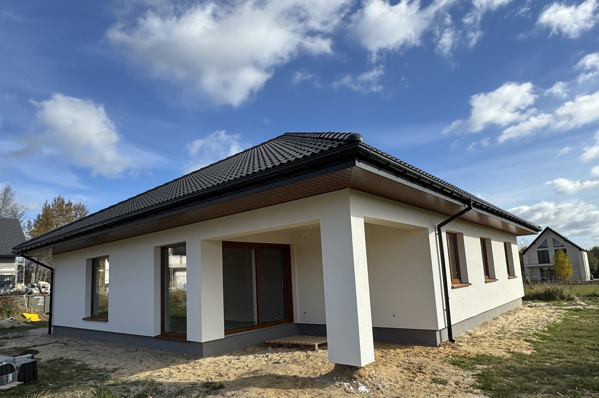 Parterowy dom modułowy z białymi ścianami i ciemną dachówką, drewniane okna, widok z zewnątrz w słoneczny dzień, teren jeszcze niezagospodarowany.