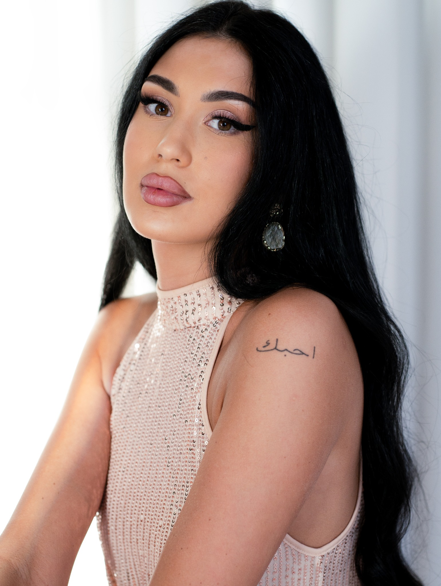 Portret kobiety o ciemnych włosach, w błyszczącej sukience, z tatuażem na ramieniu, pozującej na jasnym tle.