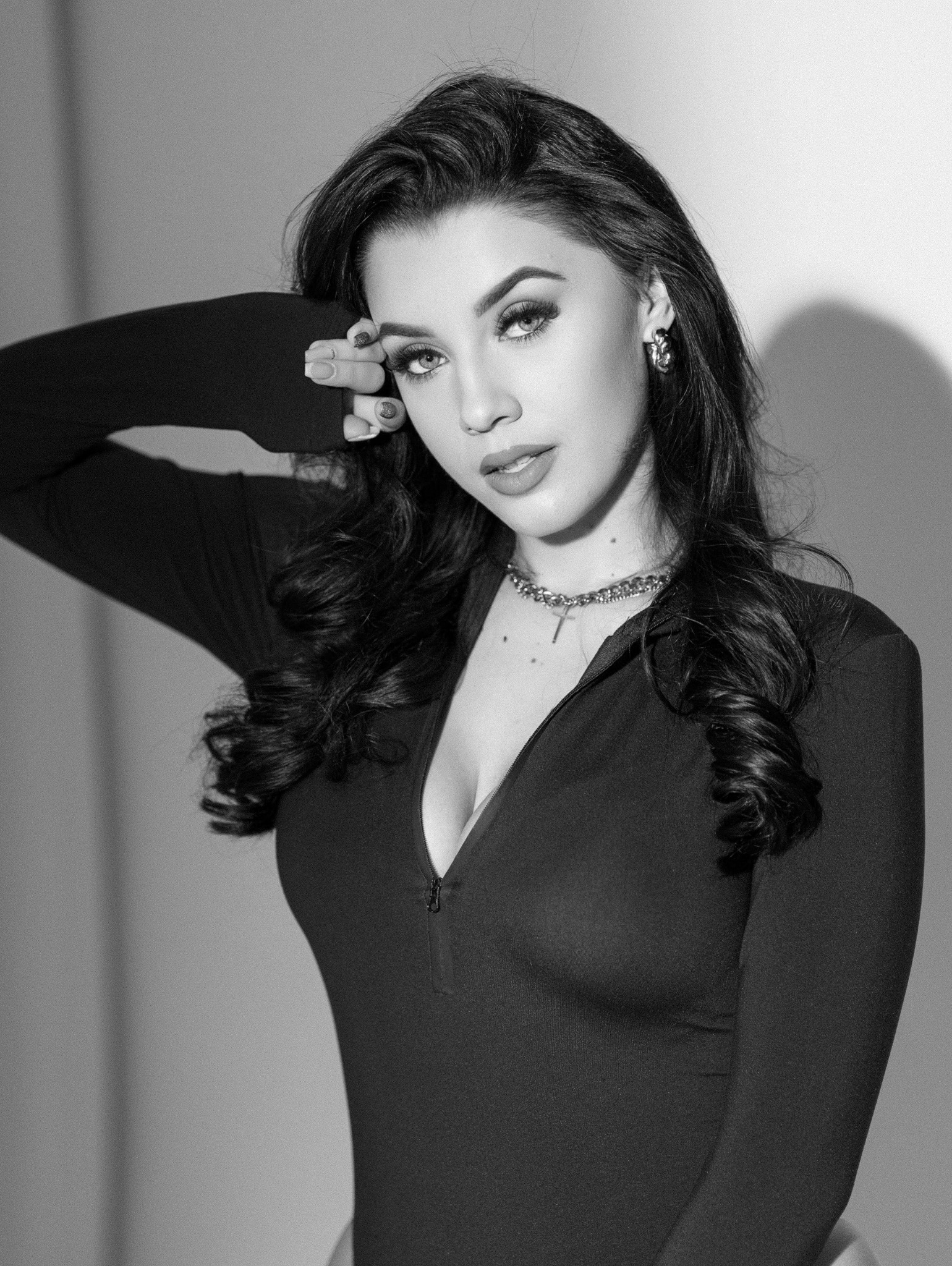 Czarno-biały portret kobiety z ciemnymi, kręconymi włosami, w dopasowanym czarnym stroju z rozpiętym zamkiem, z ręką uniesioną do twarzy, patrzącej w obiektyw.