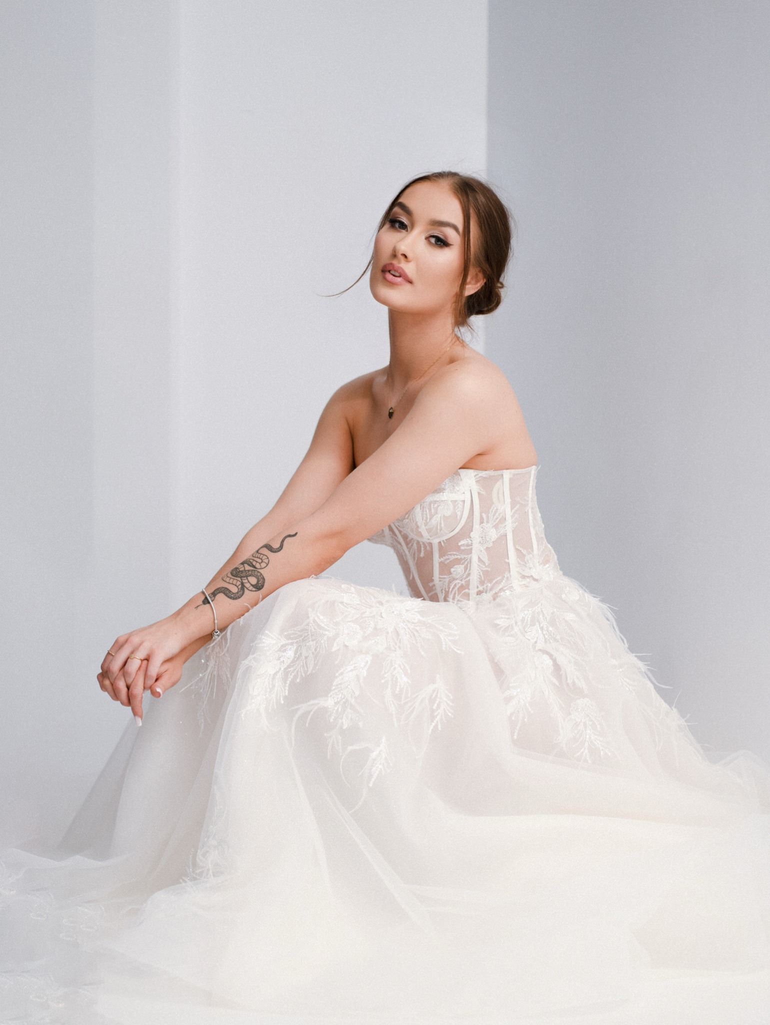 Portret kobiety w koronkowej sukni bez ramiączek, z tatuażem węża na ramieniu, siedzącej na podłodze na tle białej ściany.