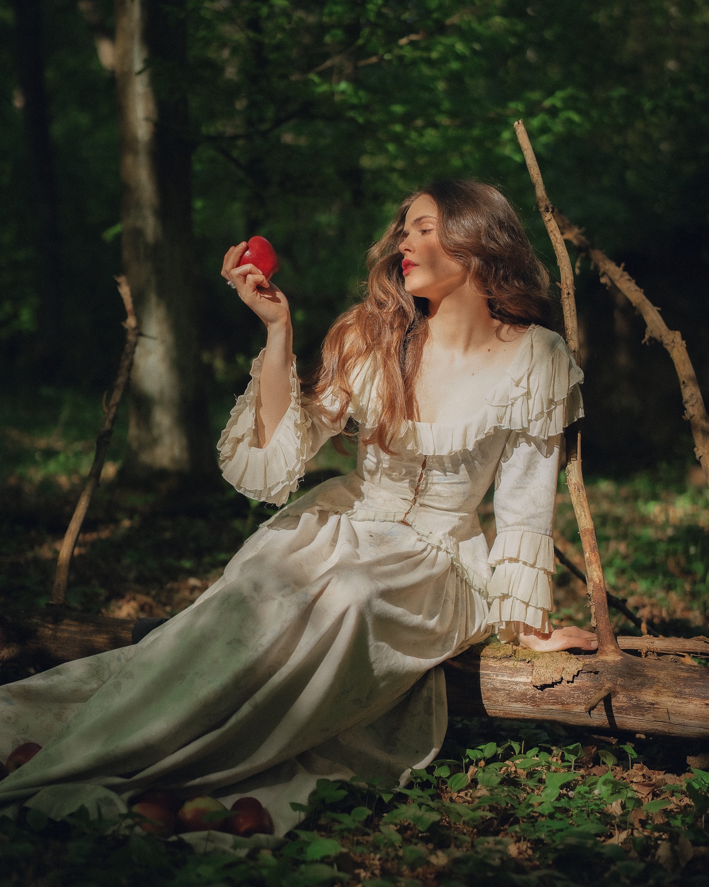 Kobieta w bieli siedzi na pniu w lesie, trzymając czerwone jabłko. Romantyczna sceneria, gra świateł i cieni, bajkowy klimat.