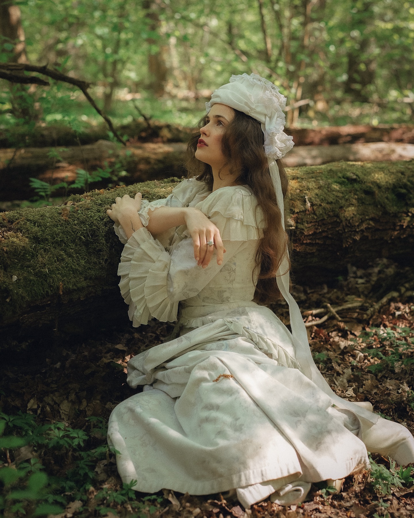 Portret kobiety w białej sukni i czepku, opierającej się o zwalone drzewo w lesie. Ujęcie w naturalnym świetle, z delikatną zielenią w tle.