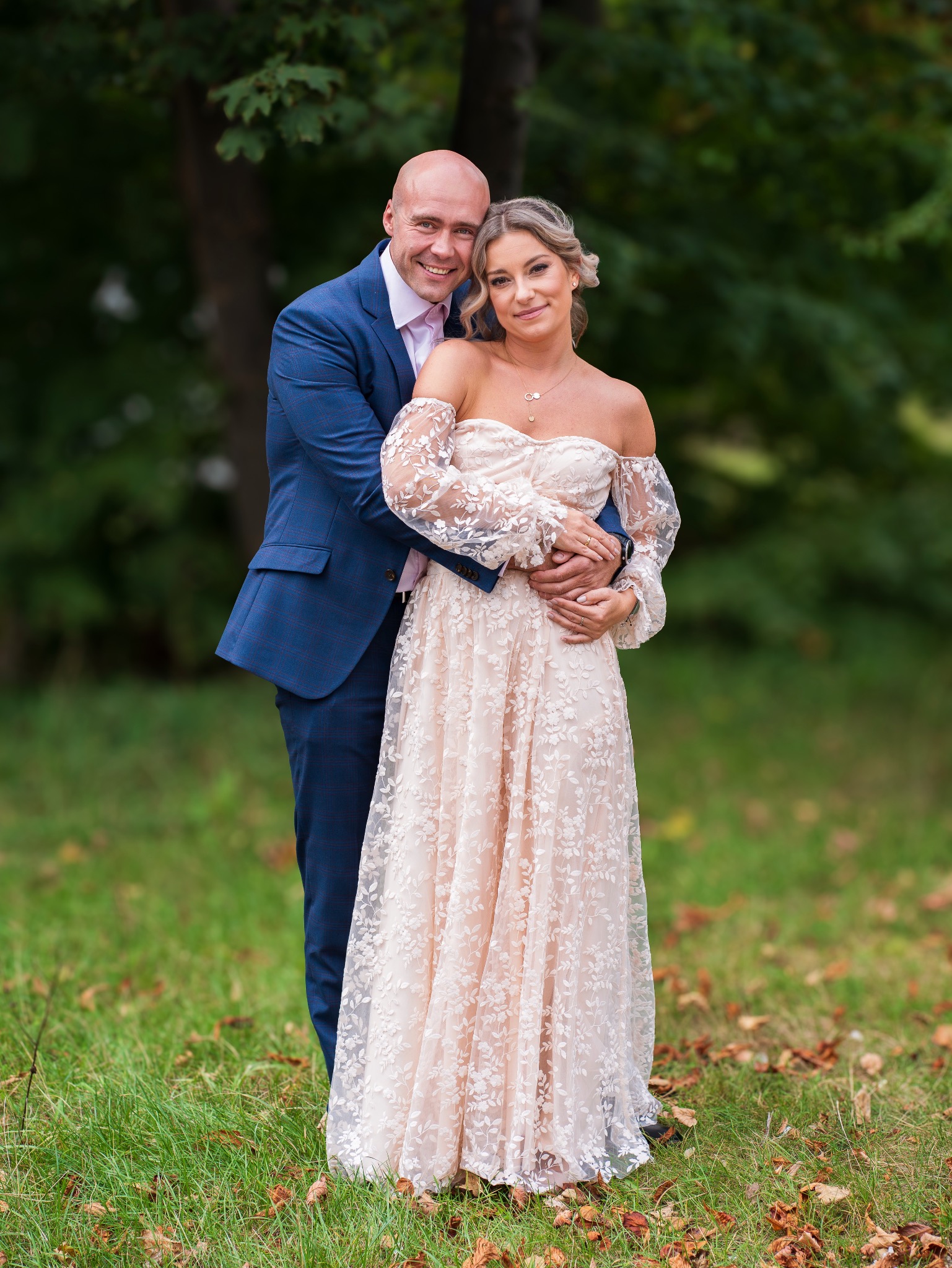 Uśmiechnięta para: mężczyzna w niebieskim garniturze obejmuje kobietę w długiej, koronkowej sukni w plenerze, z zielonym tłem drzew i trawy.