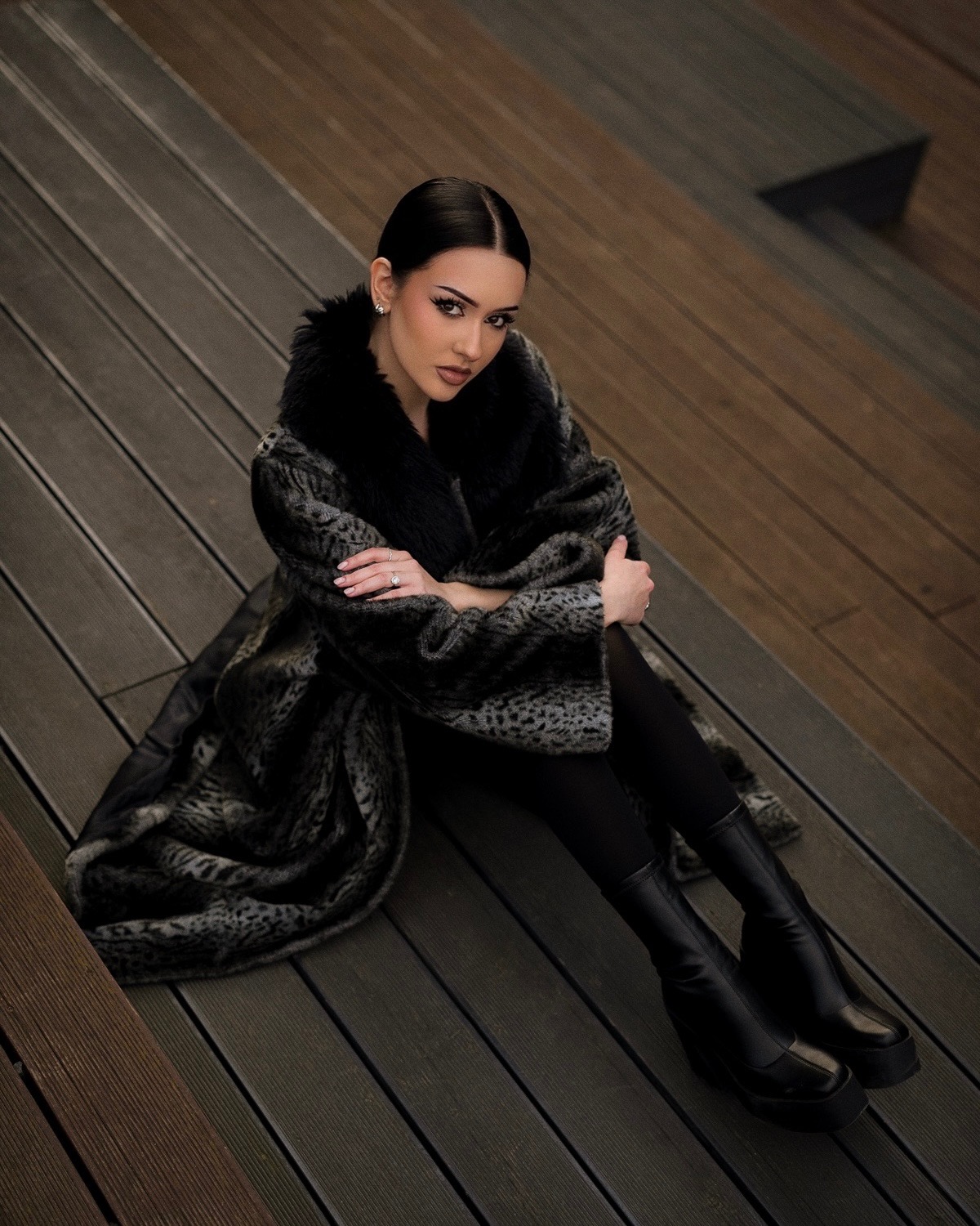 Portret kobiety w futrze z wyrazistym makijażem, siedzącej na drewnianym podeście. Ujęcie z góry, podkreślające stylizację i makijaż.