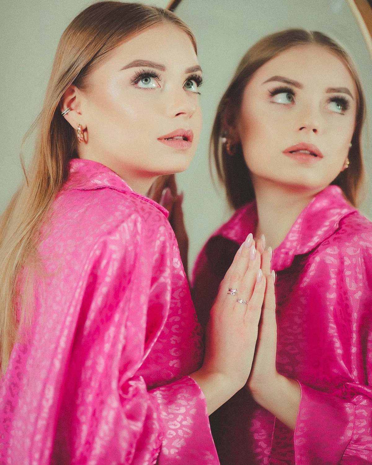 Portret młodej kobiety w różowej satynowej koszuli, odbijającej się w lustrze, z delikatnym makijażem i subtelną biżuterią. Uwagę przyciągają długie rzęsy i jasne oczy.