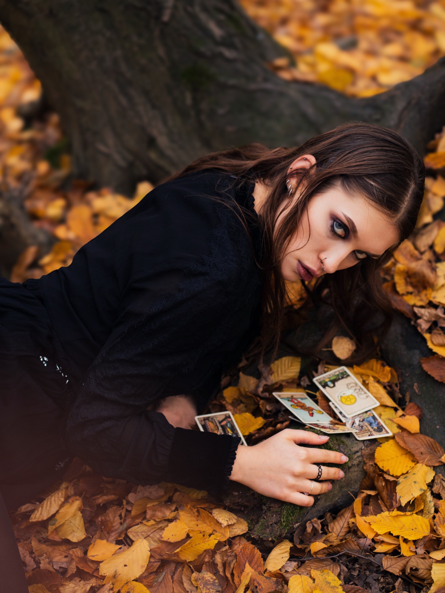Jesienna sesja portretowa: Kobieta w czerni leży na liściach z kartami tarota, intensywny makijaż oczu, piercing, artystyczne ujęcie pod drzewem, melancholijny nastrój.