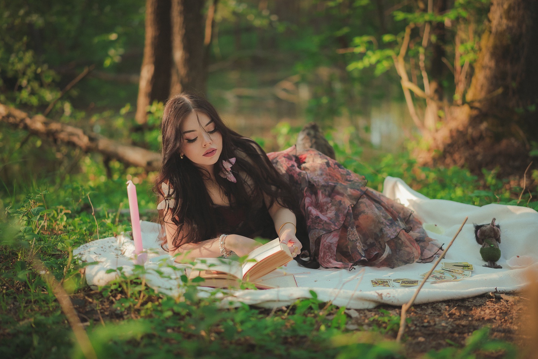Kobieta w kwiecistej sukni leży na kocu w lesie i czyta książkę. Obok świeca, figurka i karty tarota. Delikatne, rozproszone światło podkreśla magiczną atmosferę.