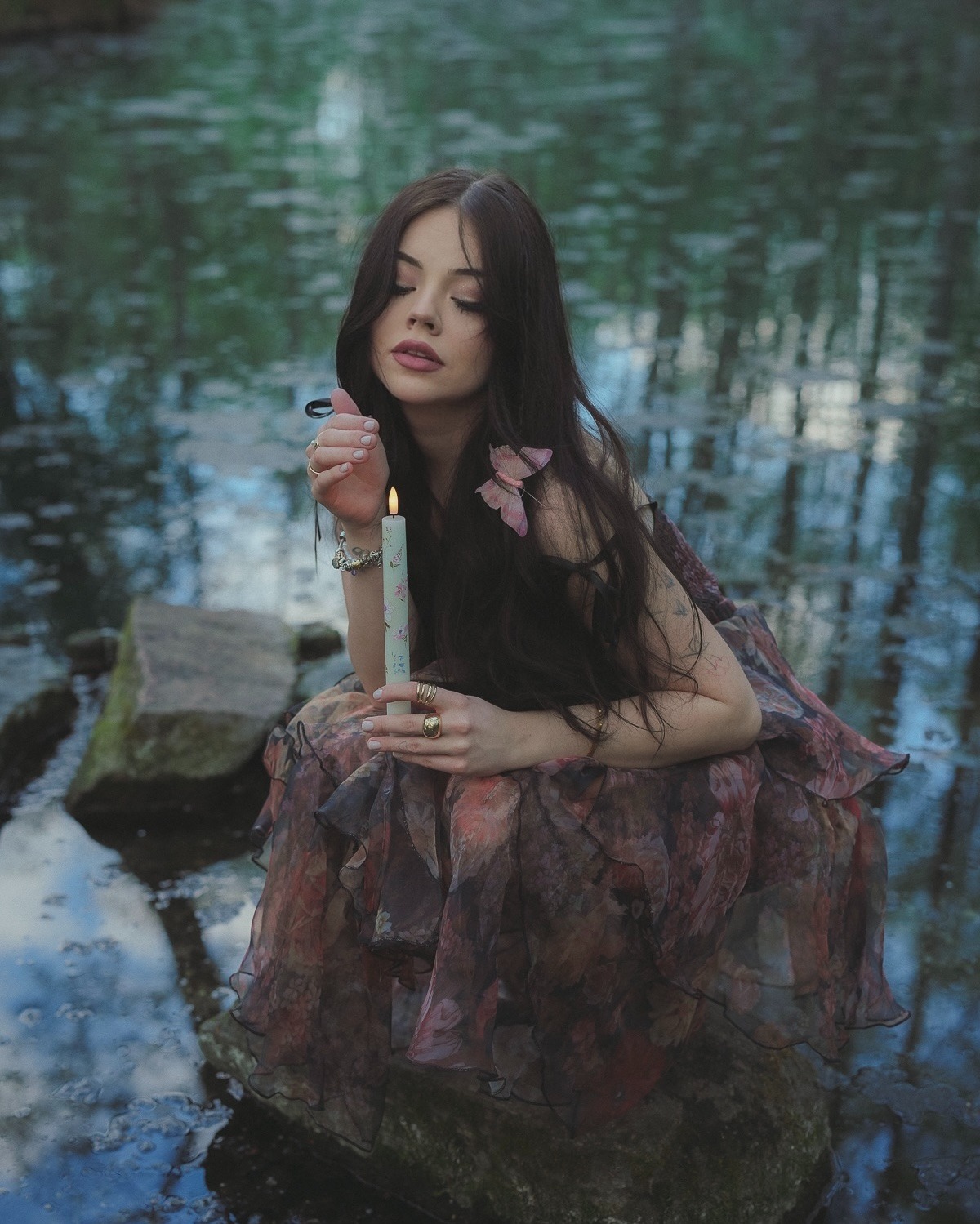 Zamyślona brunetka w kwiecistej sukience siedzi na kamieniu w wodzie, trzymając świecę. Romantyczna sesja w plenerze, w tle odbicia drzew w tafli wody.