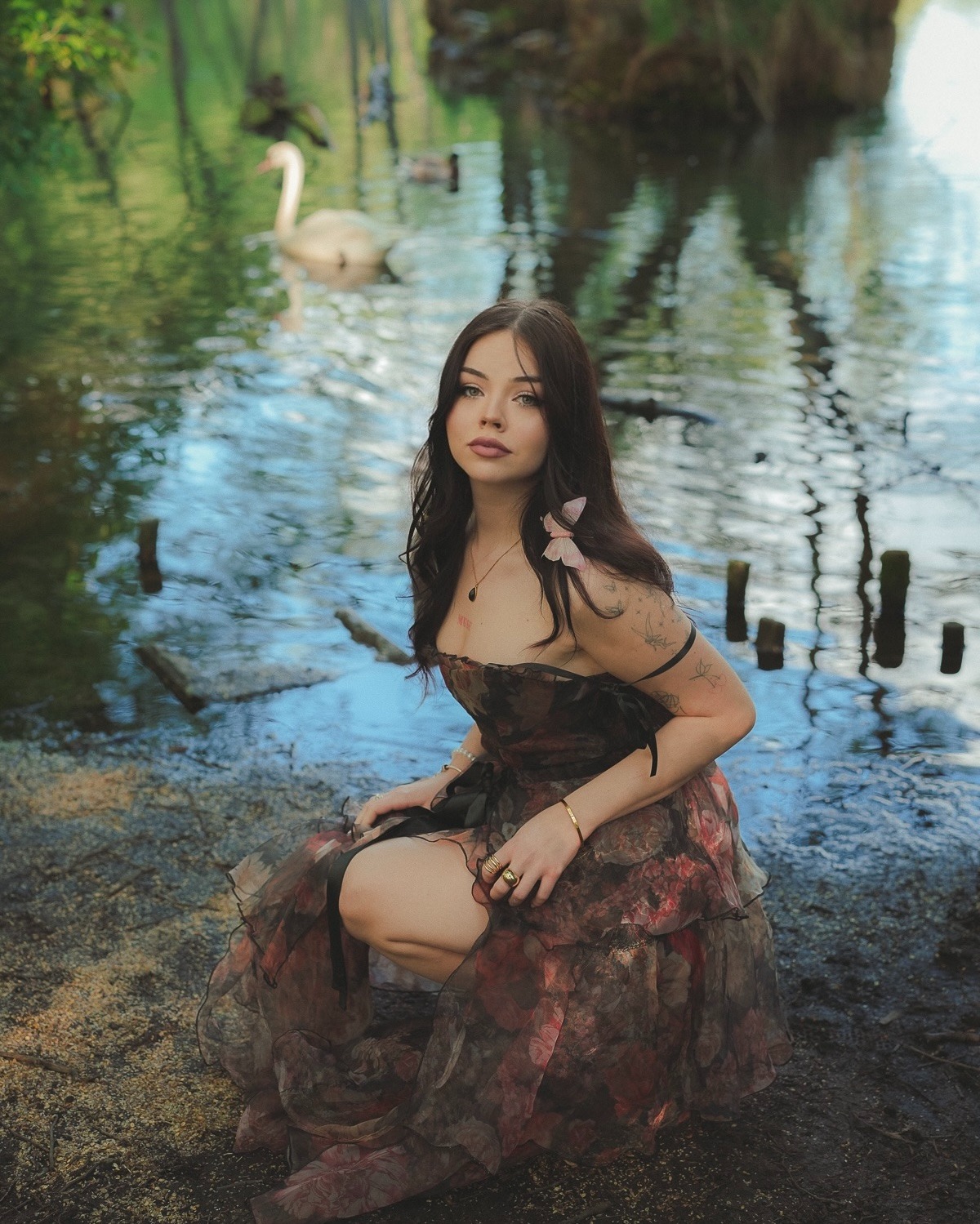 Kobieta w kwiecistej sukni klęczy na brzegu jeziora, z łabędziem w tle. Romantyczna sesja w plenerze, miękkie światło, naturalne otoczenie.