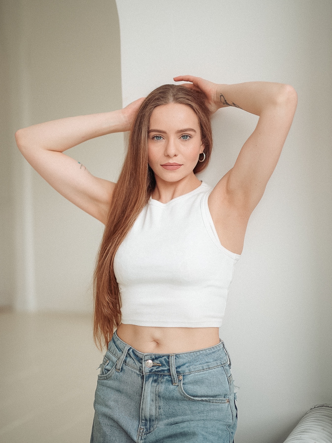 Portret kobiety w białym topie i jeansach, z rękami uniesionymi nad głową, na jasnym tle. Widoczny tatuaż na ramieniu, subtelny makijaż i srebrne kolczyki koła.