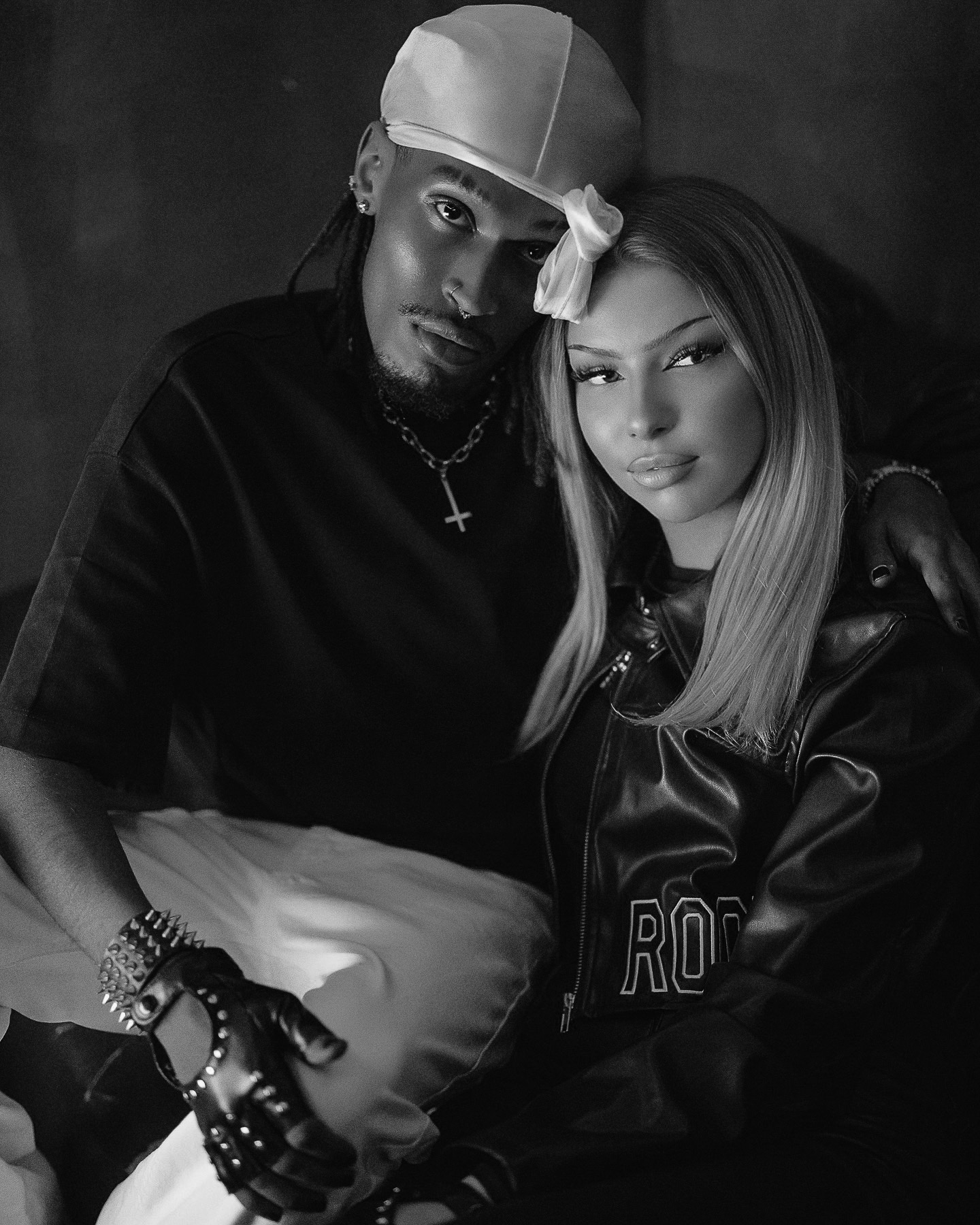 Czarno-białe, studyjne zdjęcie pary: mężczyzna z dredami w durag'u i kobieta w skórzanej kurtce z napisem 'ROCK', pozują blisko siebie, tworząc intymną atmosferę.