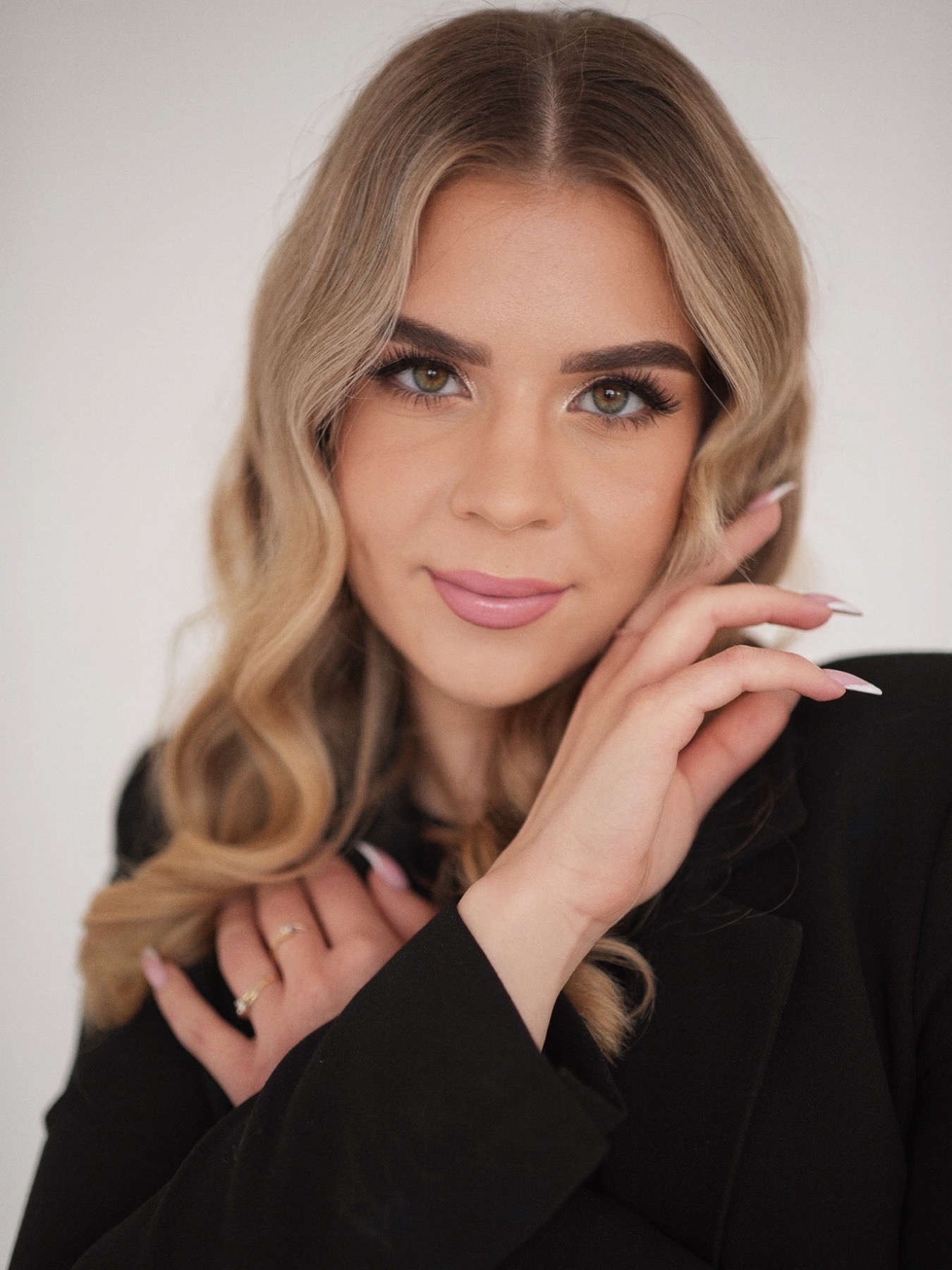 Portret kobiety z blond włosami w czarnej marynarce, delikatny makijaż, dłonie blisko twarzy, subtelny uśmiech, pierścionek na palcu, jasne tło.