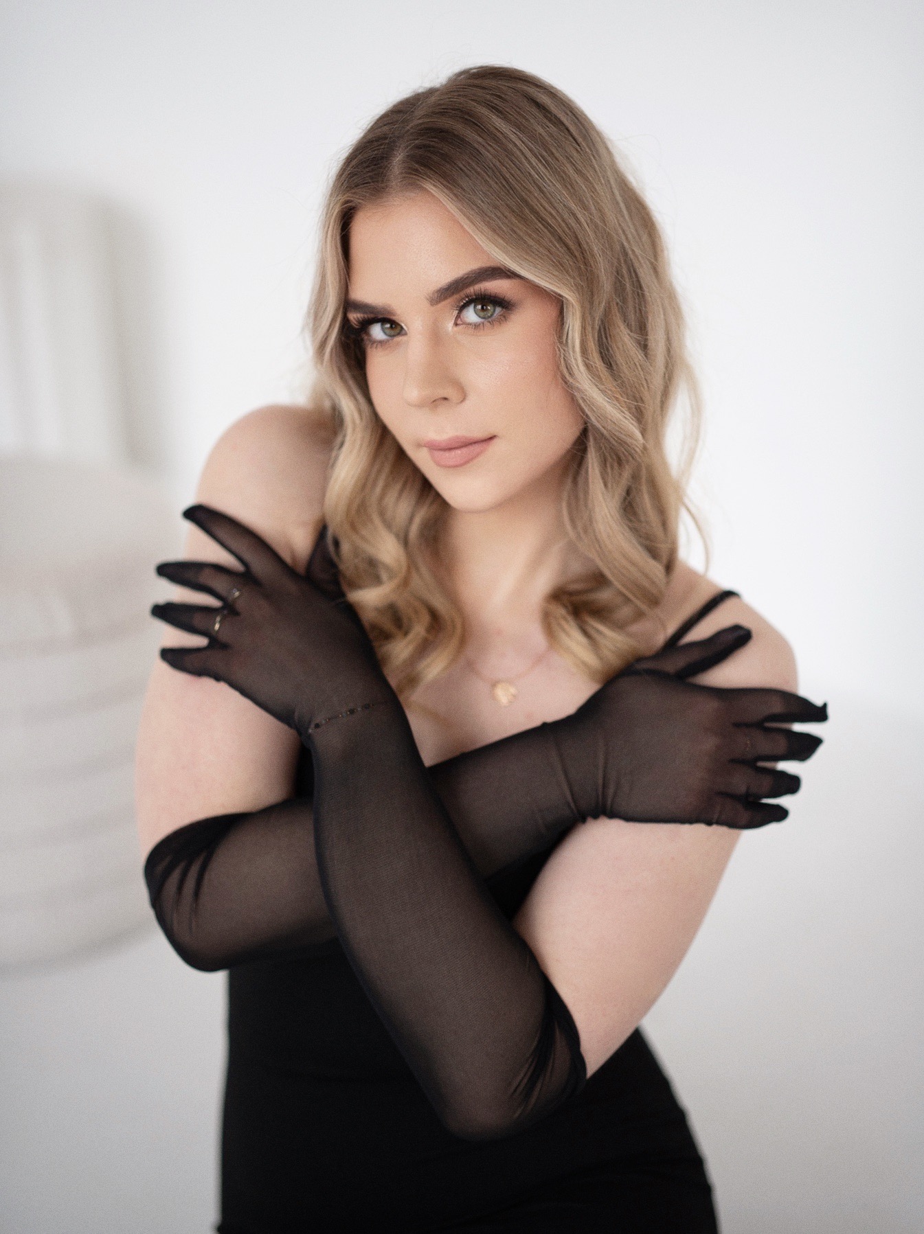 Portret kobiety w czarnej sukience i długich, przezroczystych rękawiczkach, z delikatnym makijażem i blond włosami, na jasnym tle.