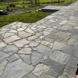 Kamienna ścieżka z nieregularnych płyt, w tle dom i krajobraz. Widoczna betoniarka sugeruje trwające prace wykończeniowe.