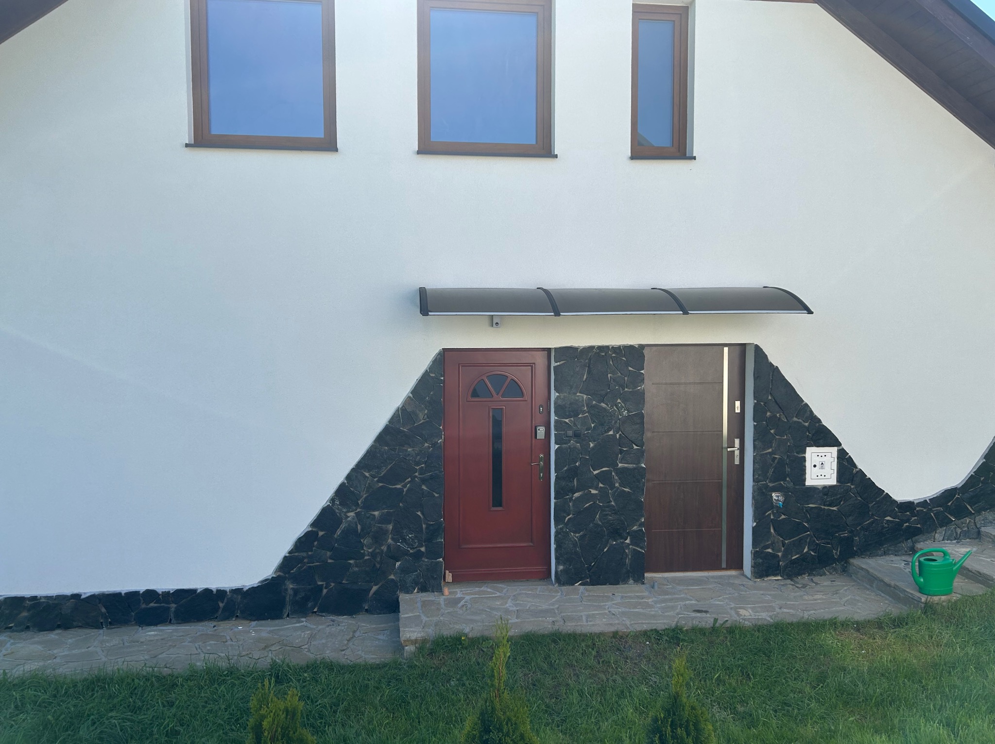 Elewacja domu z białym tynkiem i kamienną okładziną wokół wejść. Widoczne trzy okna i zielona konewka. Dwa wejścia pod wspólnym zadaszeniem.