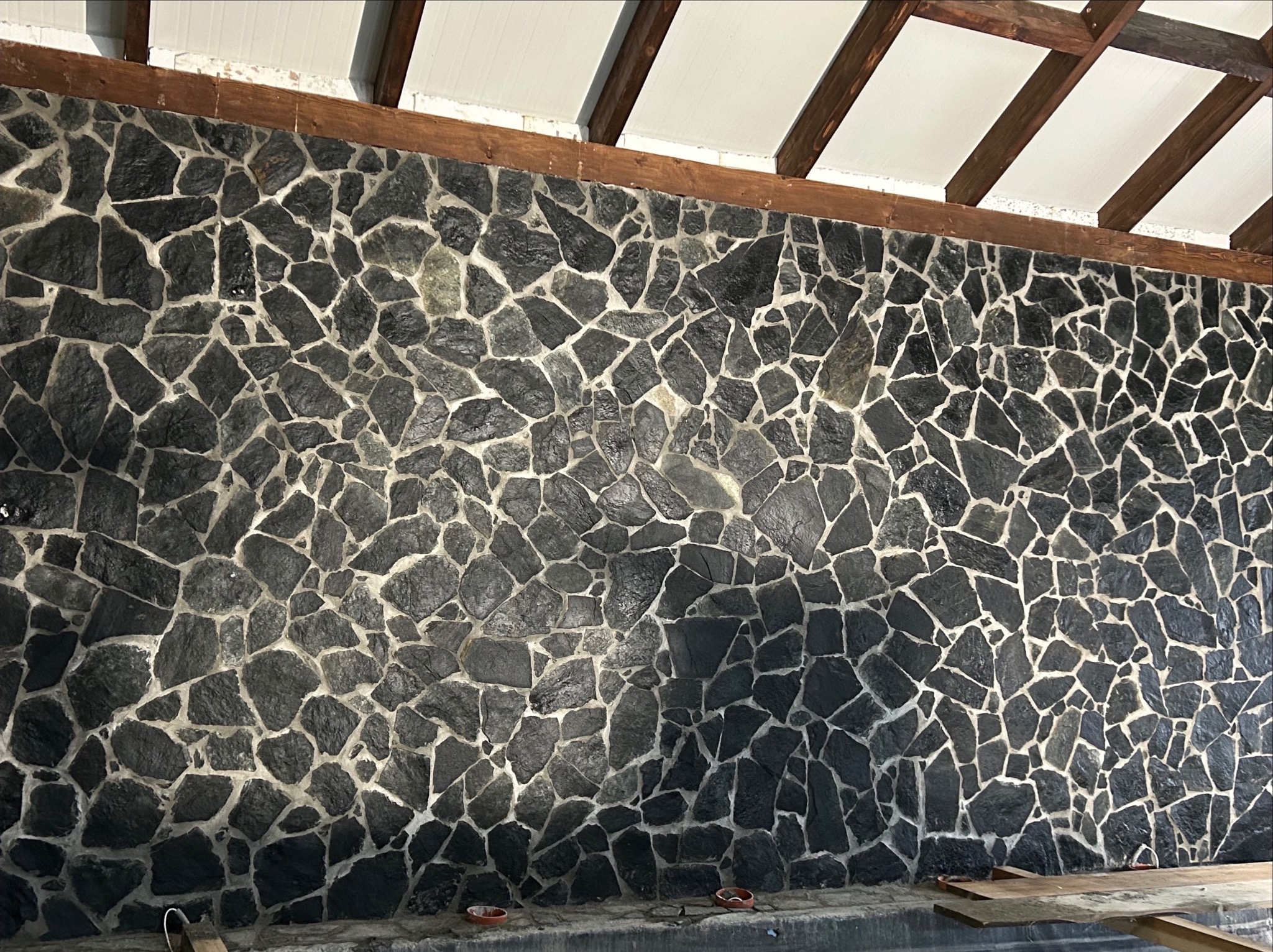 Ściana wykończona czarnym kamieniem nieregularnego kształtu z jasną fugą, drewniana konstrukcja stropu, fragment podłogi z deskami.
