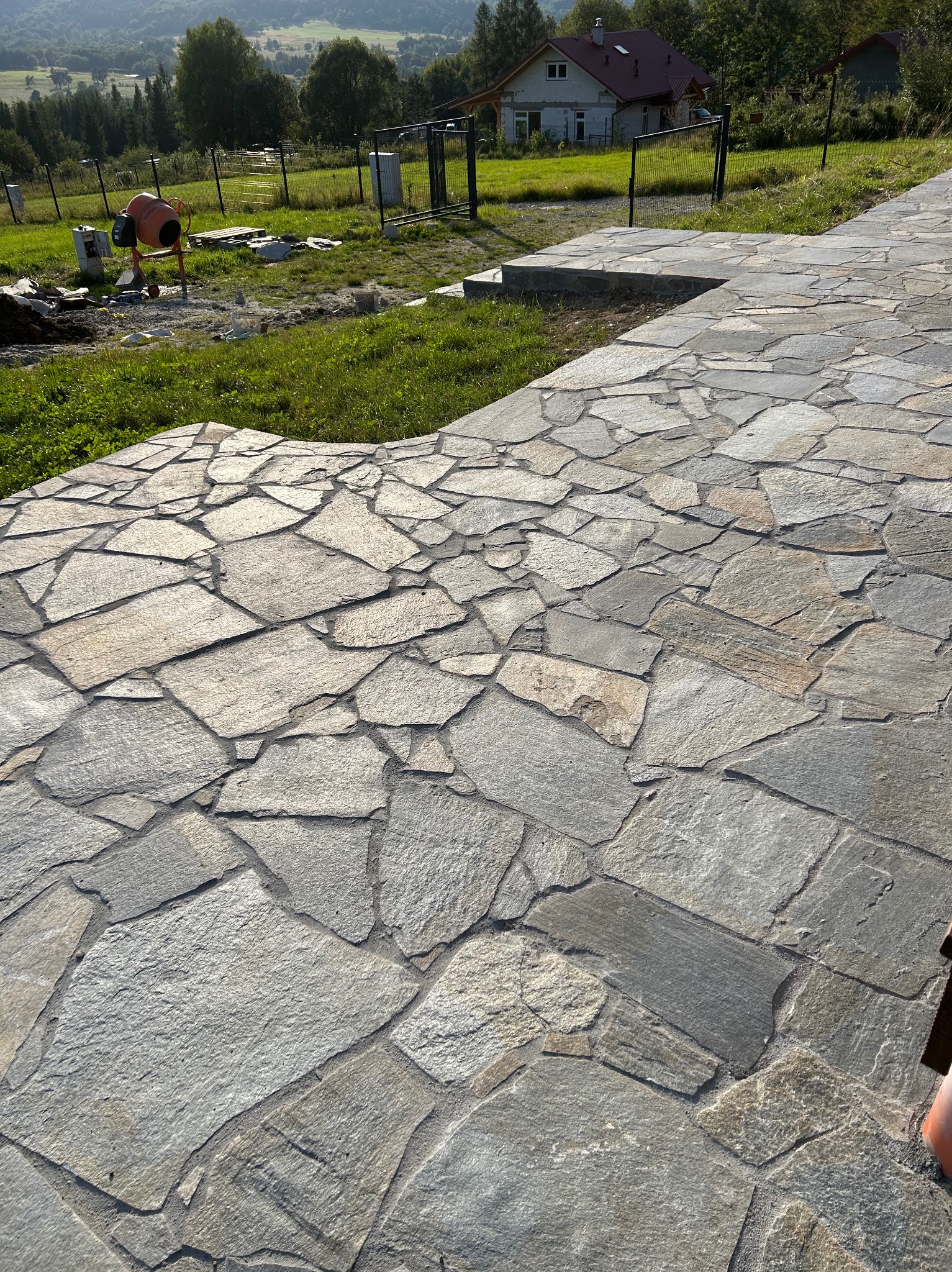 Kamienna ścieżka z nieregularnych płyt, w tle dom i krajobraz. Widoczna betoniarka sugeruje trwające prace wykończeniowe.