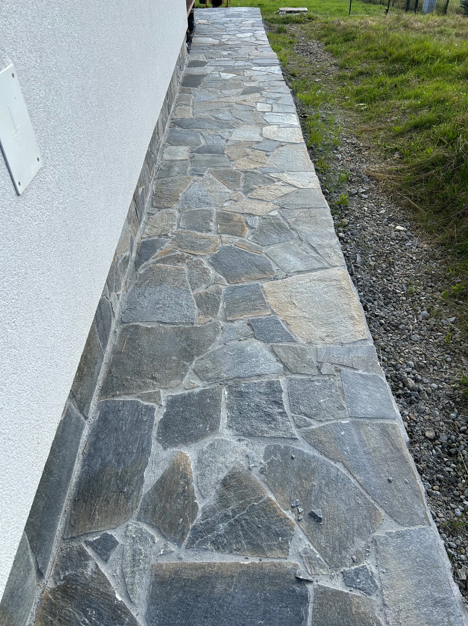 Kamienny chodnik z nieregularnych płyt, przylegający do białej ściany budynku. Widoczne trawa i kamyczki. Ujęcie z góry, naturalne światło.