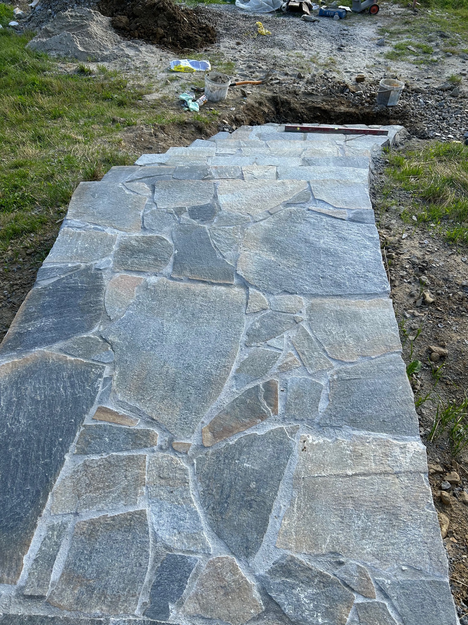 Kamienna ścieżka ułożona z nieregularnych płyt, widoczny poziomica i materiały budowlane w tle. Realizacja z kamienia naturalnego na zewnątrz.