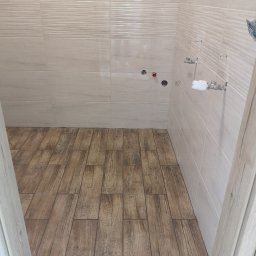 Marlux Mariusz Wójcik - Wykończona łazienka z beżowymi płytkami ściennymi i podłogą imitującą drewno. Widoczne przyłącza wodne i fragment ościeżnicy drzwi.