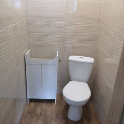 Marlux Mariusz Wójcik - Jasna łazienka po remoncie: beżowe płytki ścienne z delikatnym wzorem, drewnopodobna podłoga, biała toaleta i szafka pod umywalkę bez blatu.