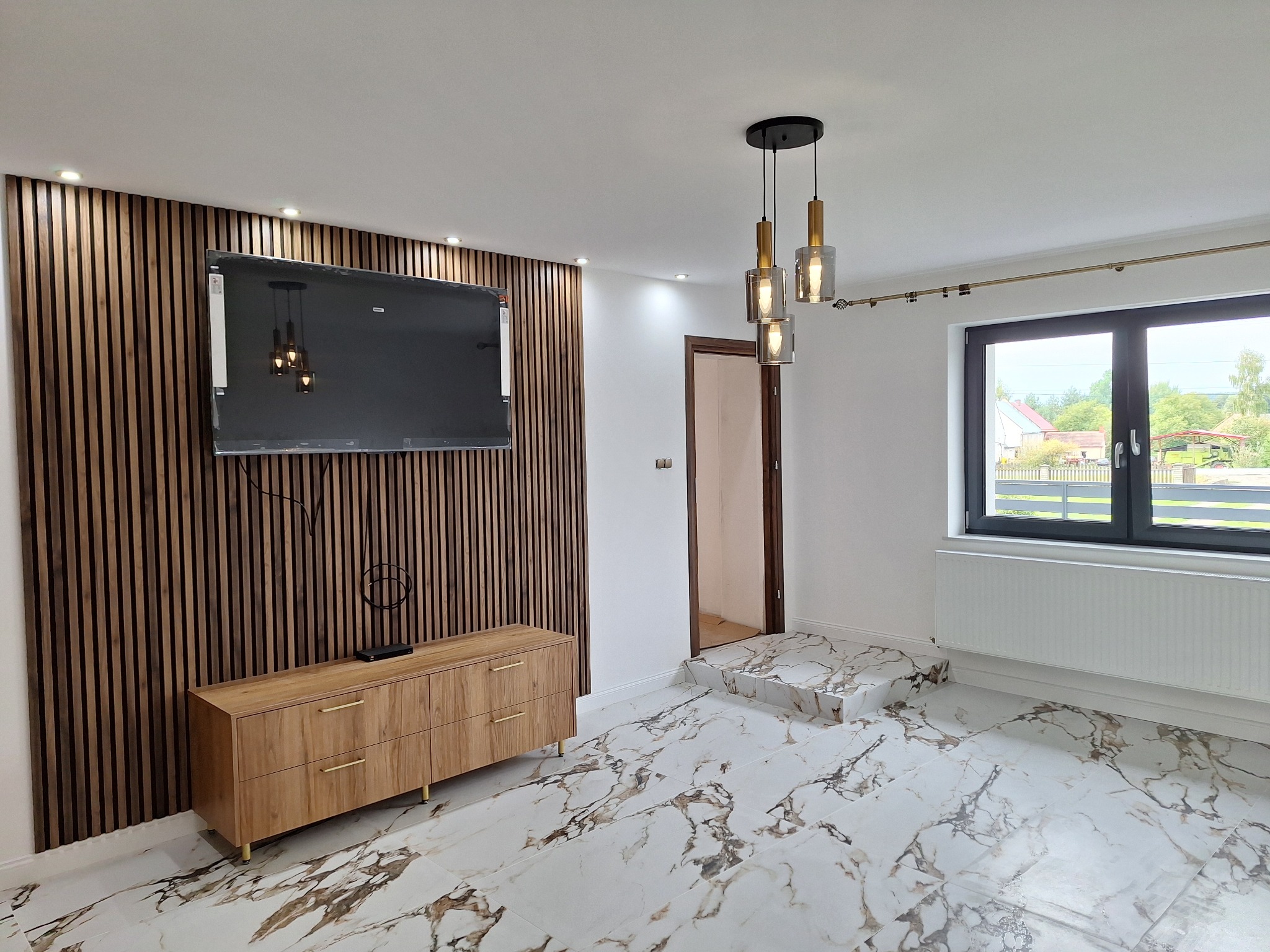 Nowoczesny salon po remoncie: marmurowe płytki, drewniana ściana z TV, komoda i lampa wisząca. Okno z widokiem na zewnątrz. Wykończenie wnętrz w jasnych barwach.