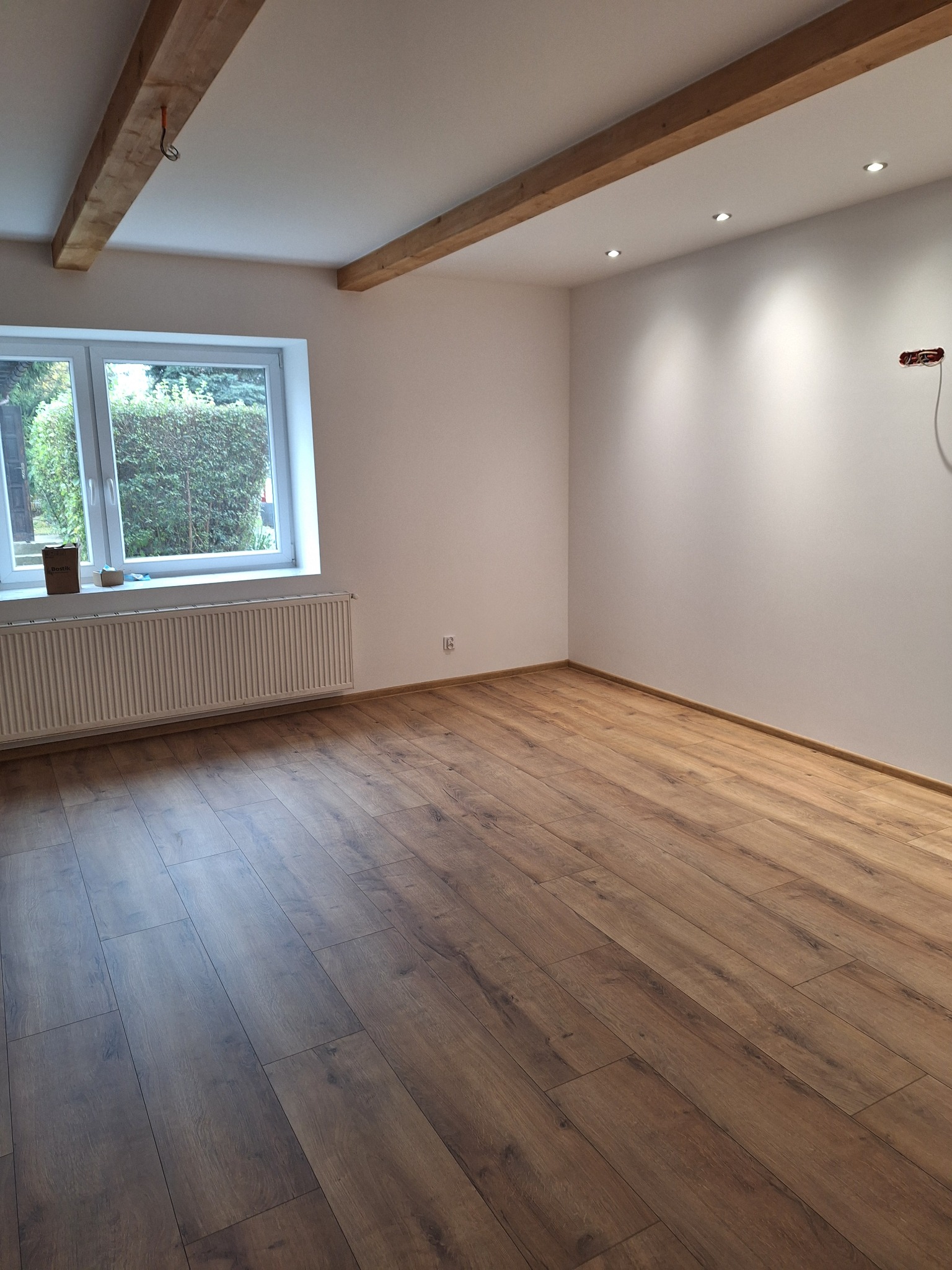 Jasne wnętrze z drewnianą podłogą i belkami stropowymi. Widoczne okno z widokiem na zieleń, grzejnik i gładkie, białe ściany z punktowym oświetleniem.