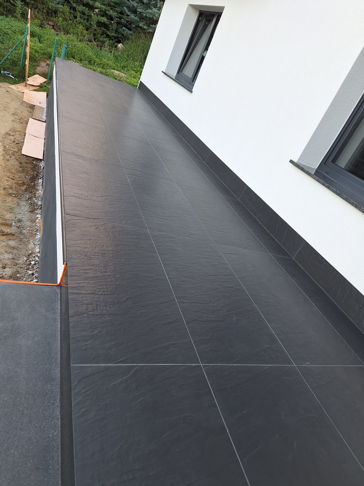 Ciemne płytki gresowe na zewnątrz budynku, ułożone równo z fugami. Widoczne okna z szarymi ramami i fragment trawnika. Minimalistyczny design.