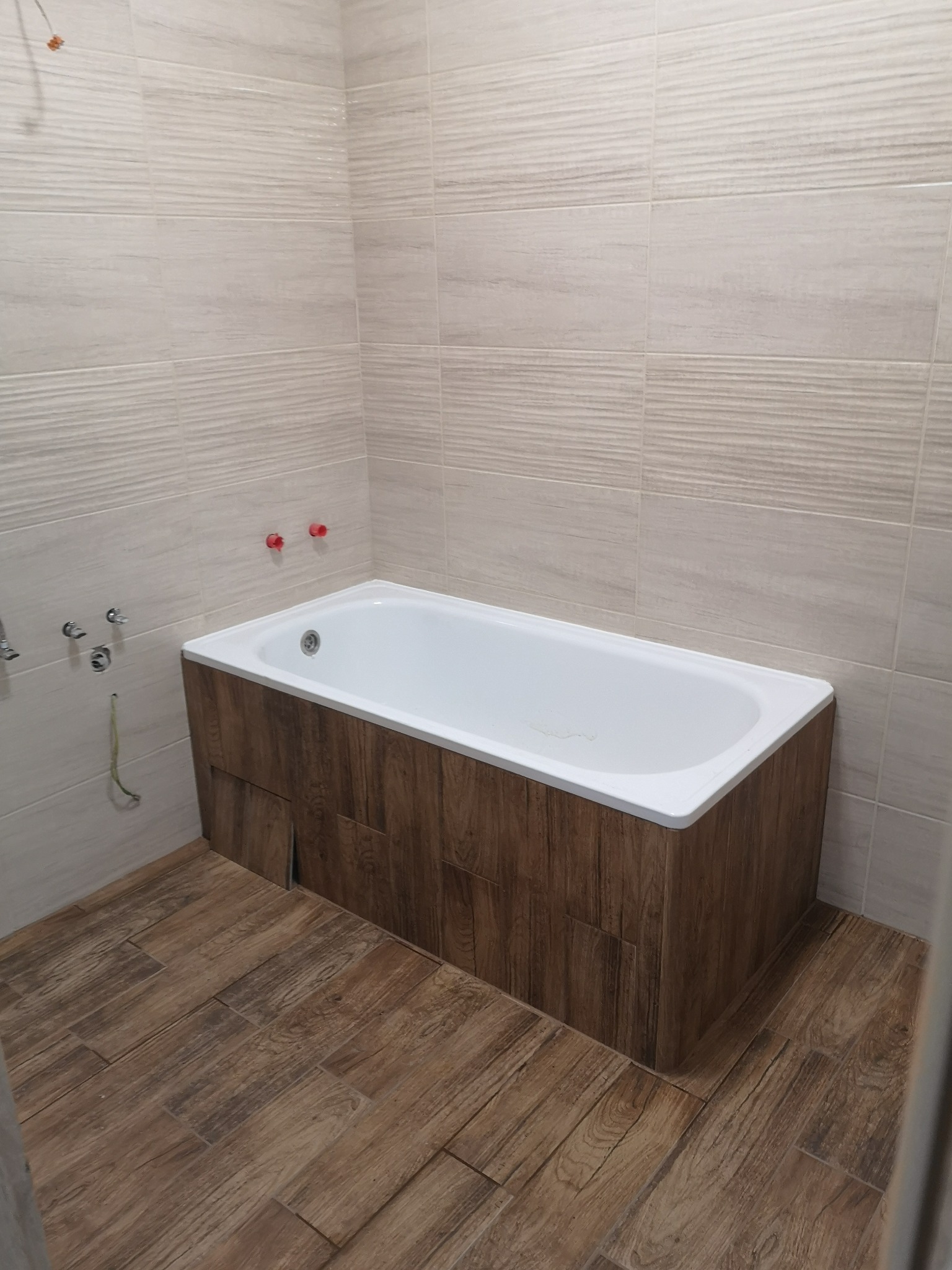 Nowoczesna łazienka z wanną obudowaną drewnopodobnymi panelami, beżowe płytki na ścianach i podłodze. Widoczne przyłącza wodne.