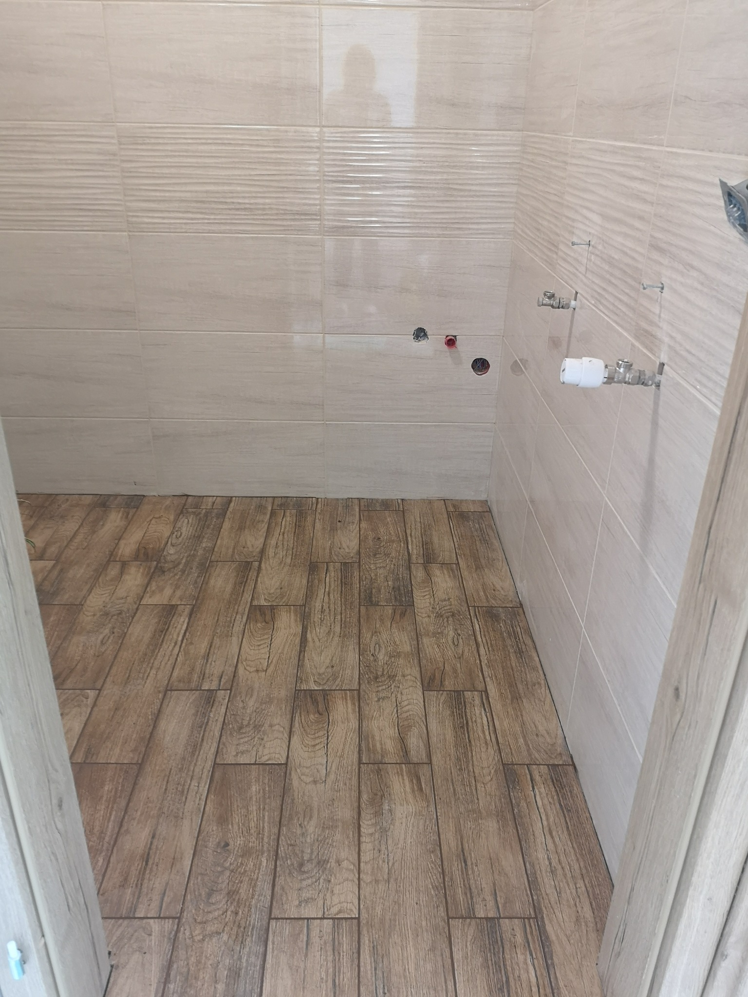 Wykończona łazienka z beżowymi płytkami ściennymi i podłogą imitującą drewno. Widoczne przyłącza wodne i fragment ościeżnicy drzwi.