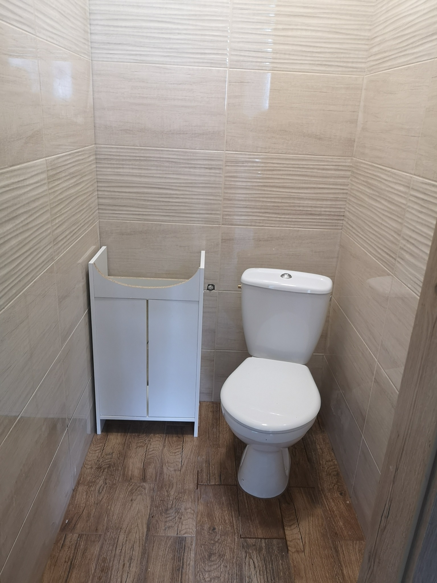 Jasna łazienka po remoncie: beżowe płytki ścienne z delikatnym wzorem, drewnopodobna podłoga, biała toaleta i szafka pod umywalkę bez blatu.