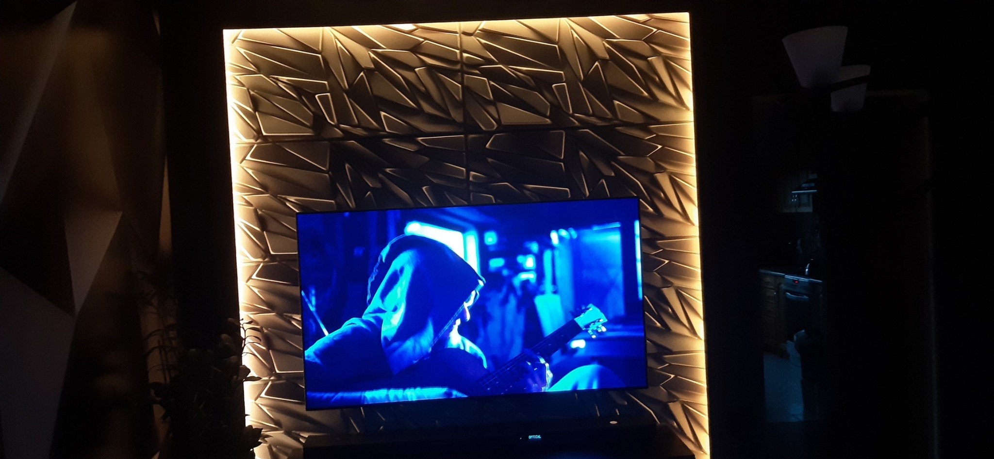 Panel dekoracyjny 3D z podświetleniem LED za telewizorem, tworzący geometryczny wzór na ścianie. Nowoczesny design wnętrza w ciemnym pomieszczeniu, z widocznym fragmentem kuchni.