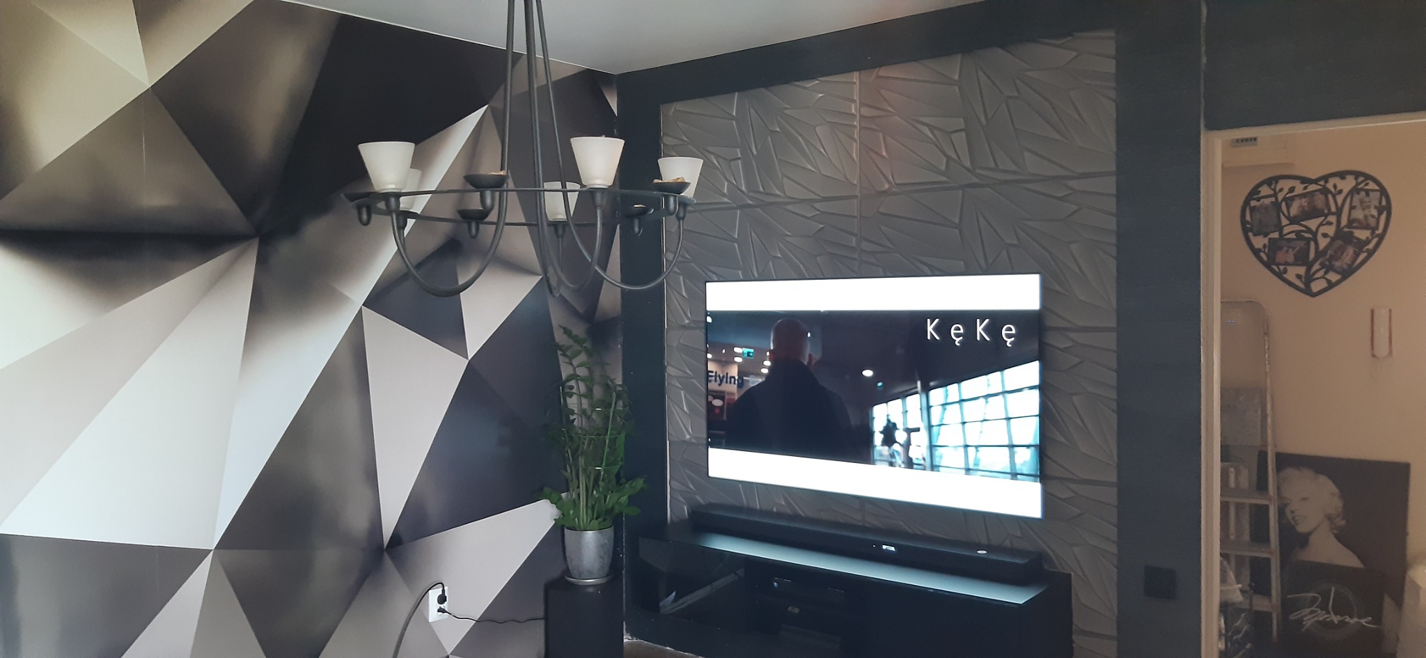 Nowoczesny salon z geometryczną tapetą, panelem 3D za telewizorem i designerskim żyrandolem. Minimalistyczny wystrój wnętrza z akcentami czerni i bieli.