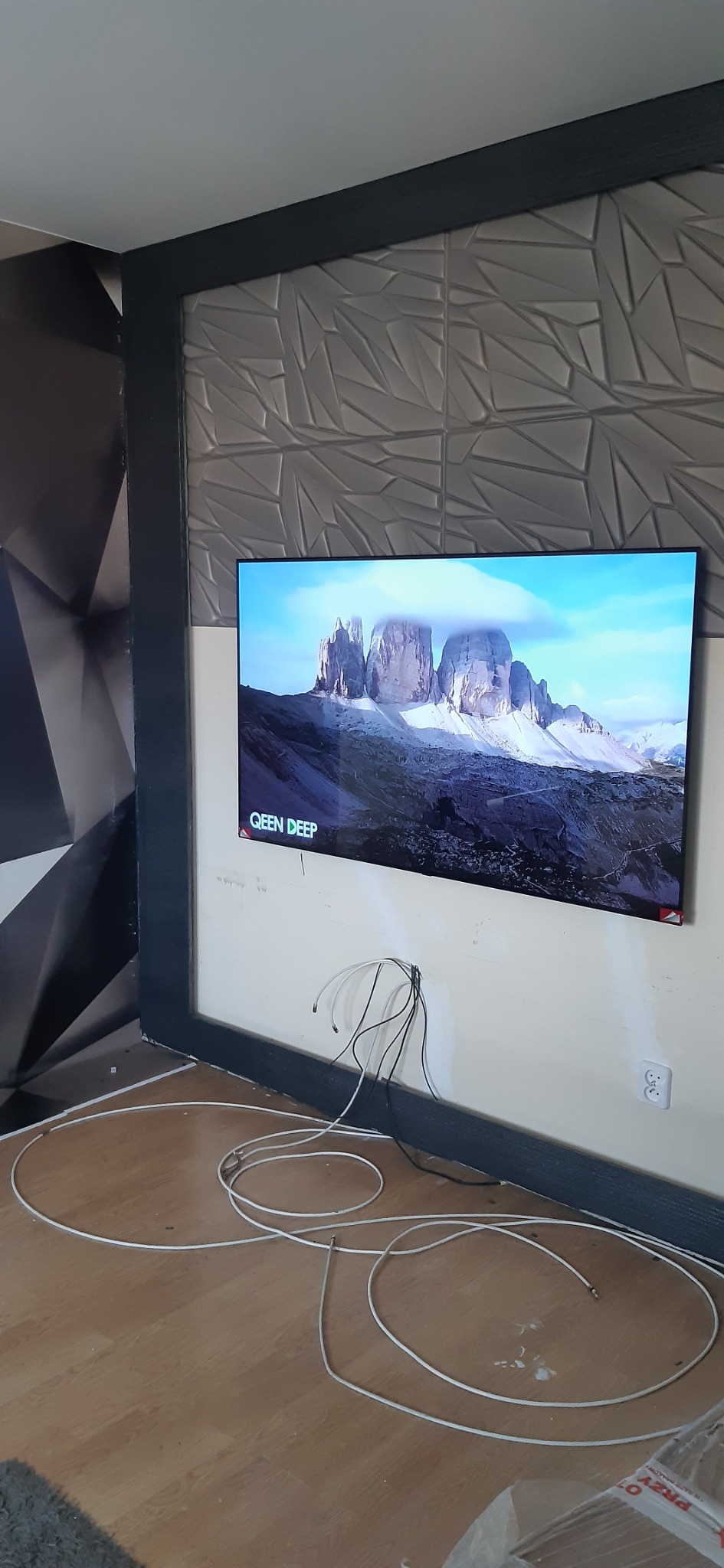 Telewizor na ścianie z panelami 3D, widoczne kable i gniazdko. Fragment drewnianej podłogi i ściany z geometrycznym wzorem. Ujęcie z dołu, brak wykończenia.