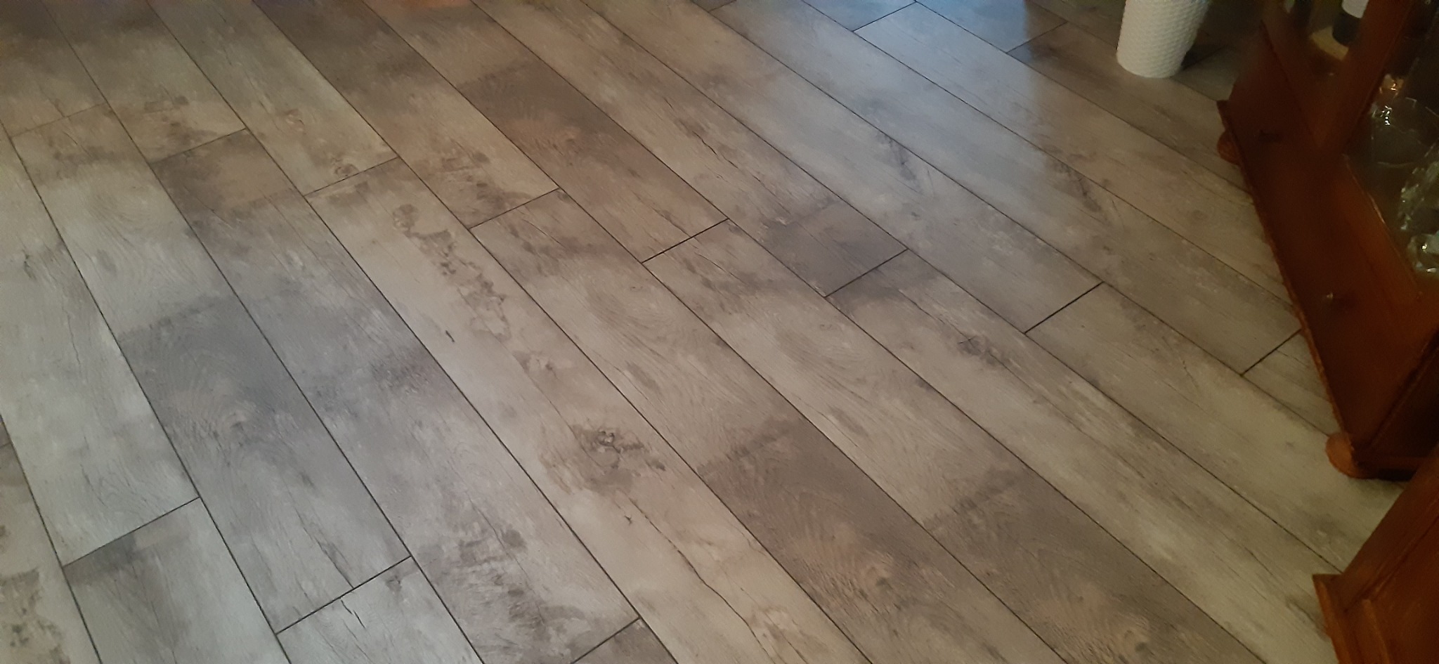 Ułożone panele podłogowe w jasnych odcieniach drewna, z widoczną strukturą słojów. Fragment drewnianej komody w prawym dolnym rogu i biała doniczka w tle.