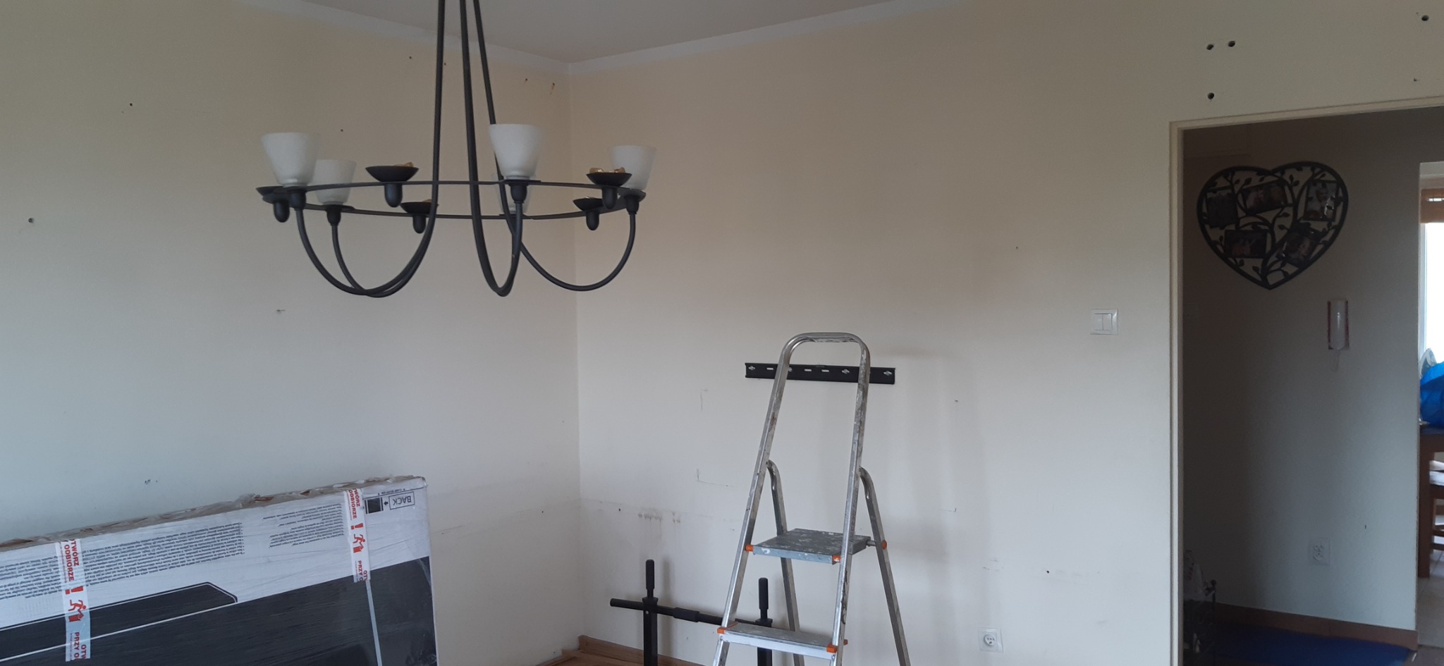 Pokój przed remontem: drabina, lampa, kartony, ślady po wierceniu w ścianach. Widoczne przygotowania do malowania lub montażu. Dekoracja w kształcie serca w przejściu.
