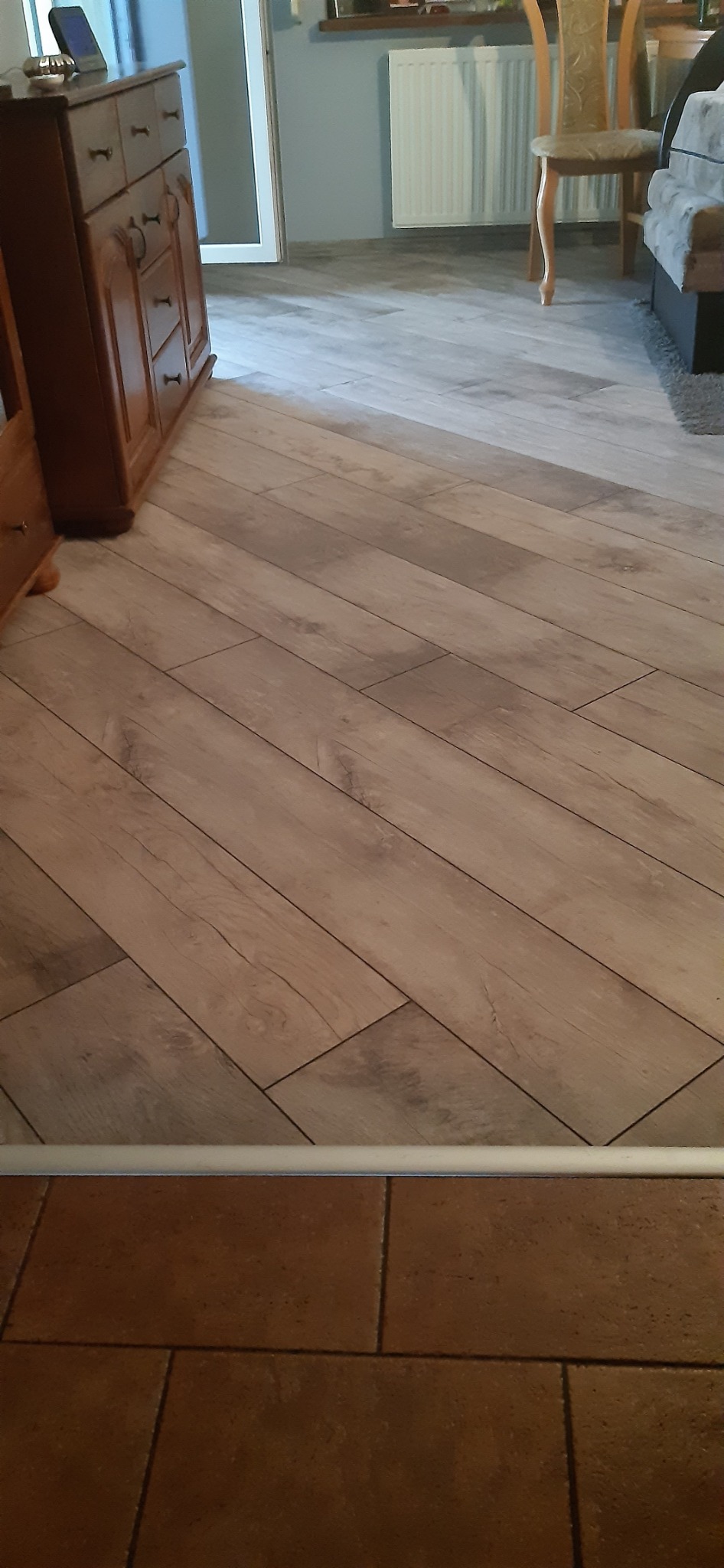 Ułożone panele podłogowe w jodełkę, widok z perspektywy podłogi, z widocznymi meblami w tle. Jasne panele z wyraźną strukturą drewna.