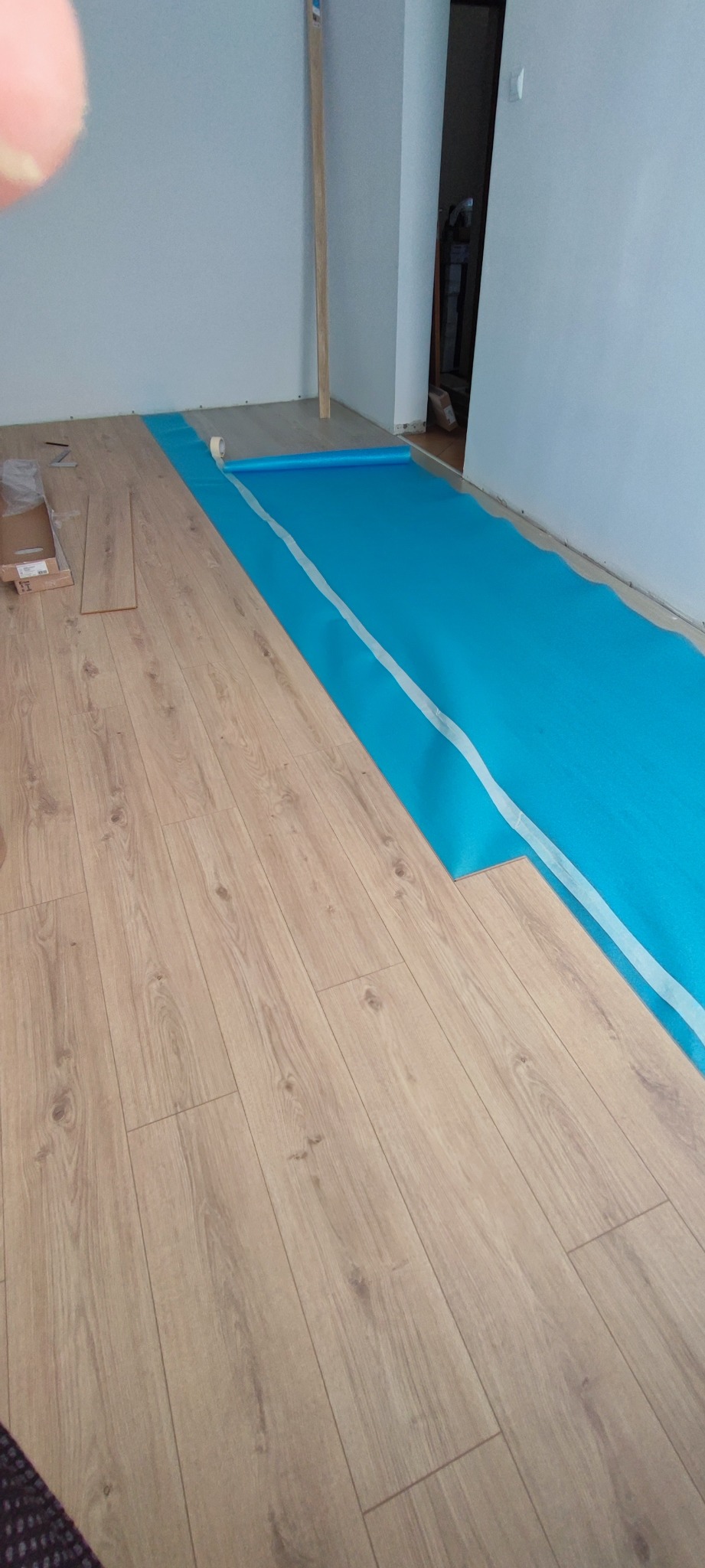 Fragment pokoju w trakcie układania paneli podłogowych. Część podłogi wyłożona panelami, reszta pokryta niebieską folią izolacyjną. Widoczne narzędzia i materiały.