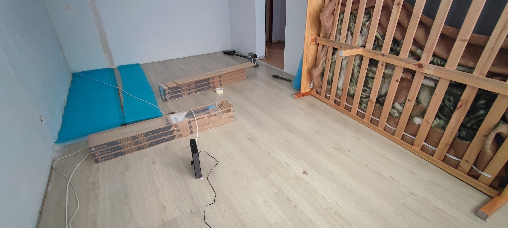 Wnętrze w trakcie układania paneli podłogowych. Widoczne paczki paneli, podkład, częściowo ułożona podłoga i narzędzia. W tle drzwi do innego pomieszczenia.