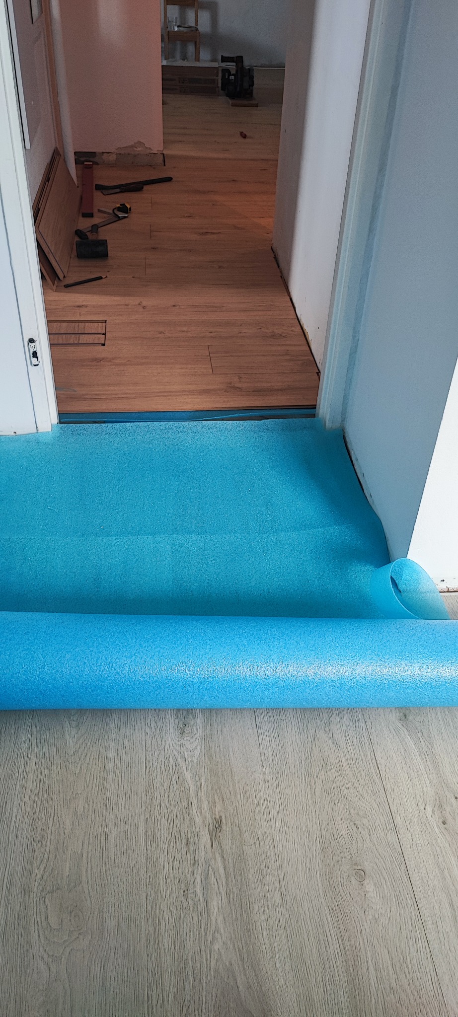 Przygotowanie do położenia paneli: podkład z niebieskiej pianki rozłożony na podłodze, widoczne panele i narzędzia w przejściu do innego pomieszczenia.