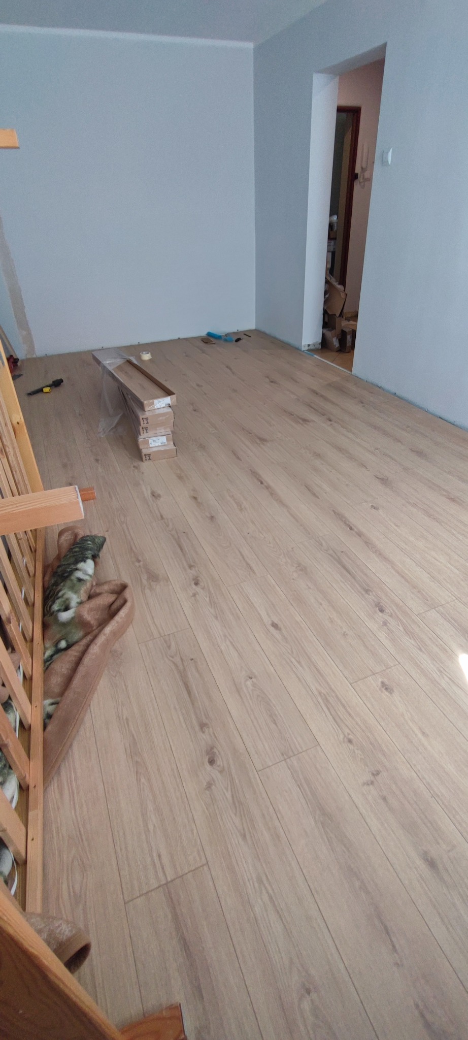 Jasne panele podłogowe ułożone w pokoju z białą ścianą. Częściowo widoczne narzędzia i paczki paneli. Widok na próg drzwiowy.