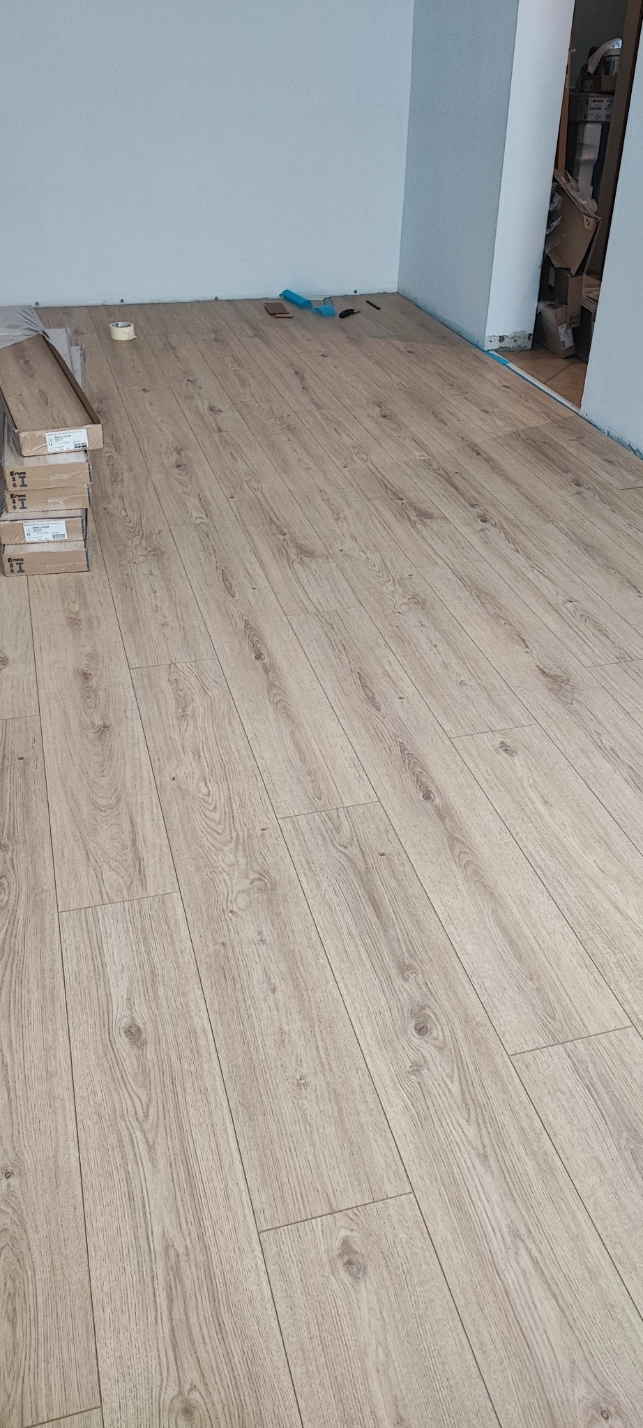 Ułożone panele podłogowe w jasnym odcieniu drewna, z widocznymi paczkami paneli i narzędziami w rogu pomieszczenia. Widok na nową podłogę po montażu.