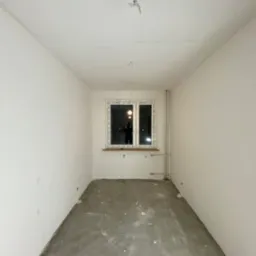 Pusty pokój po wstępnych pracach: białe ściany, surowy beton na podłodze z widocznymi śladami po remoncie, okno z widokiem na nocne miasto, przygotowane instalacje elektryczne i grzewcze.